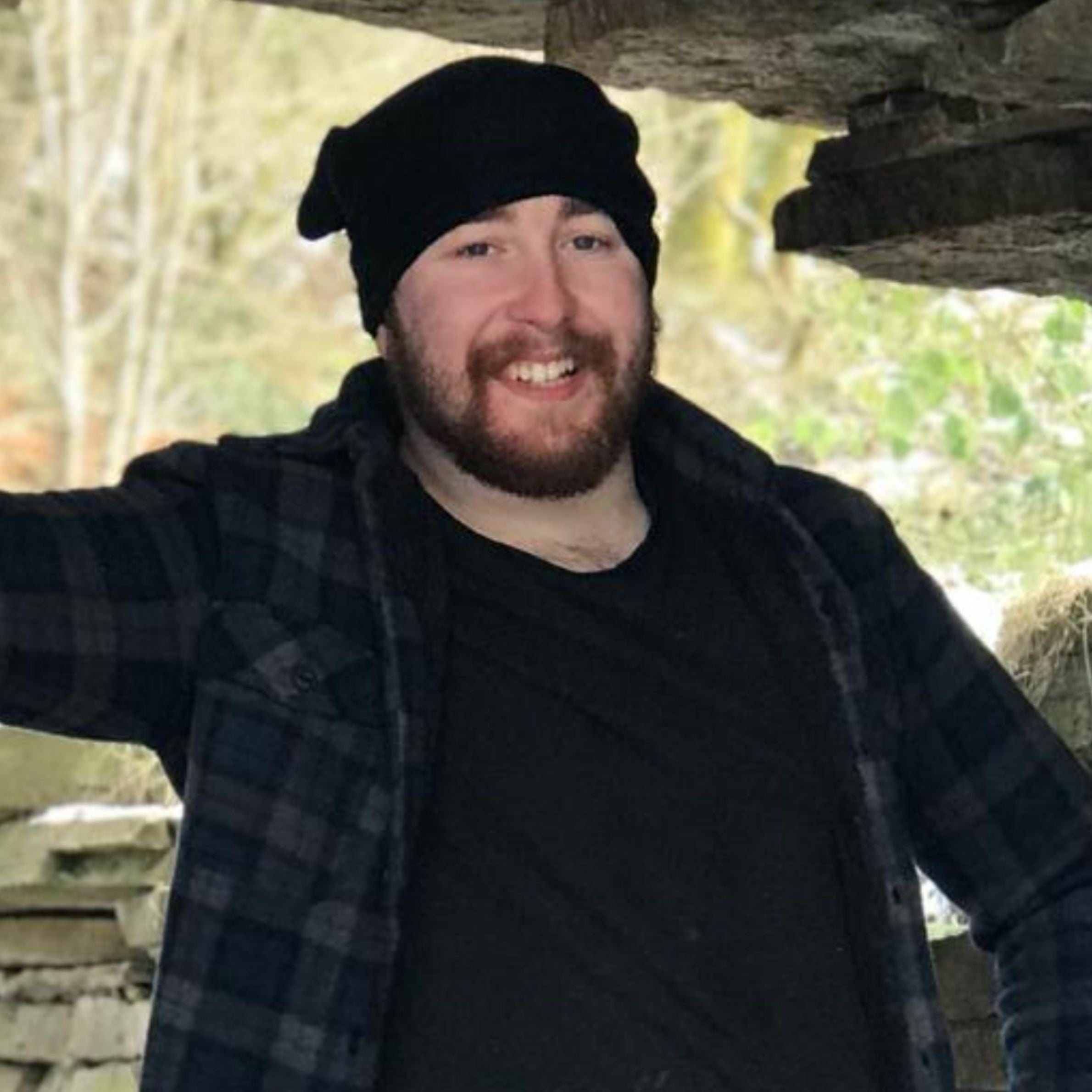 man wearing a black t shirt with a dark blue checkered shirt on top. he is also wearing a black beanie hat. he has a dark beard and is smiling for a photo. there is trees and rocks behind him
