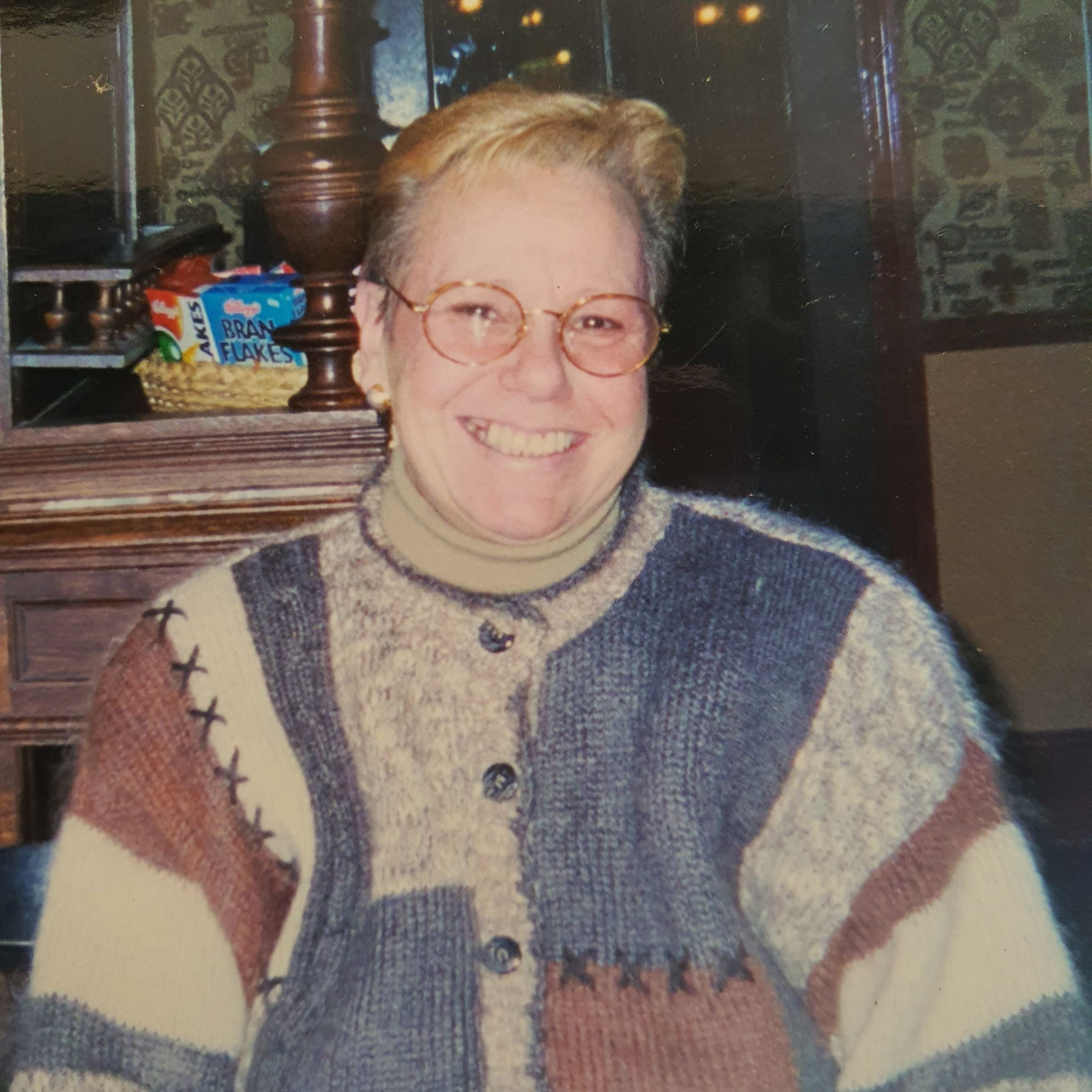 picture of a woman wearing a knitted cardigan with blue, red and grey squares on it. she is smiling and wearing glasses and has short blonde hair. there is a wooden mantle in the background with red and blue cereal boxes on it