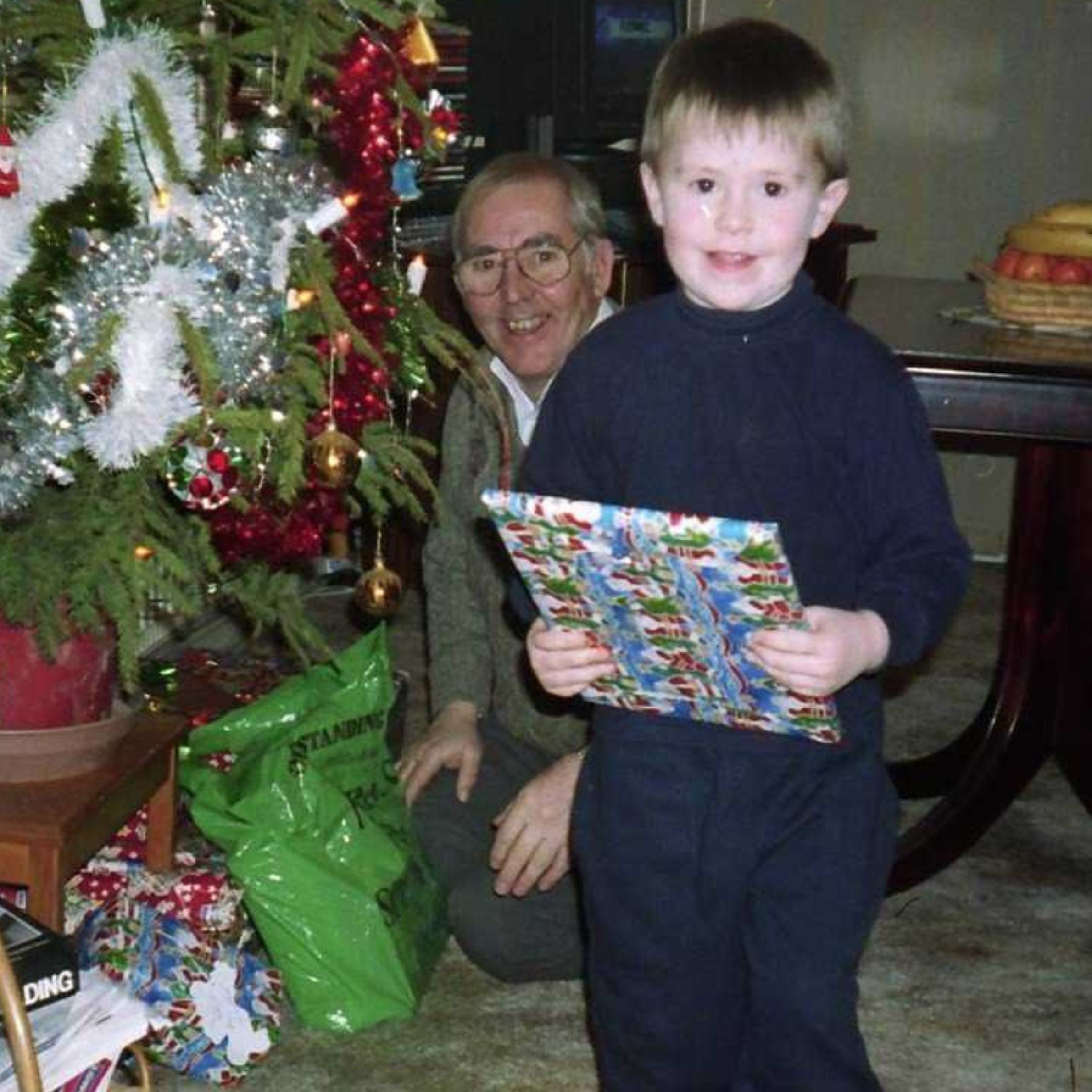 Little boy in blue top holding Christmas present and an old man behind him smiling. Theres a Christmas tree with red and white tinsel and gifts underneath it.