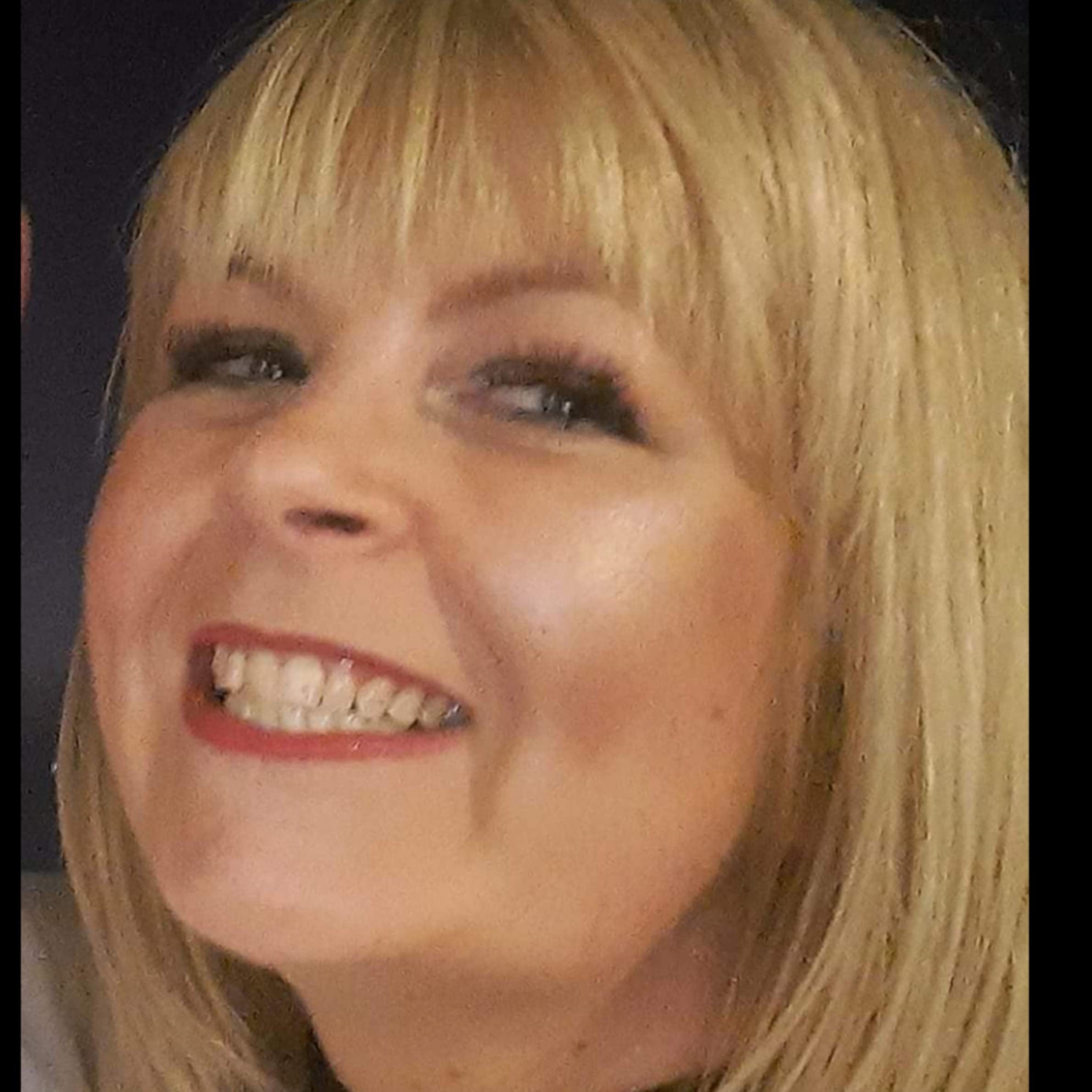 Lady with blonde straight hair and fringe smiling at the camera. She is wearing red lipstick and has dark eyeshadow on.