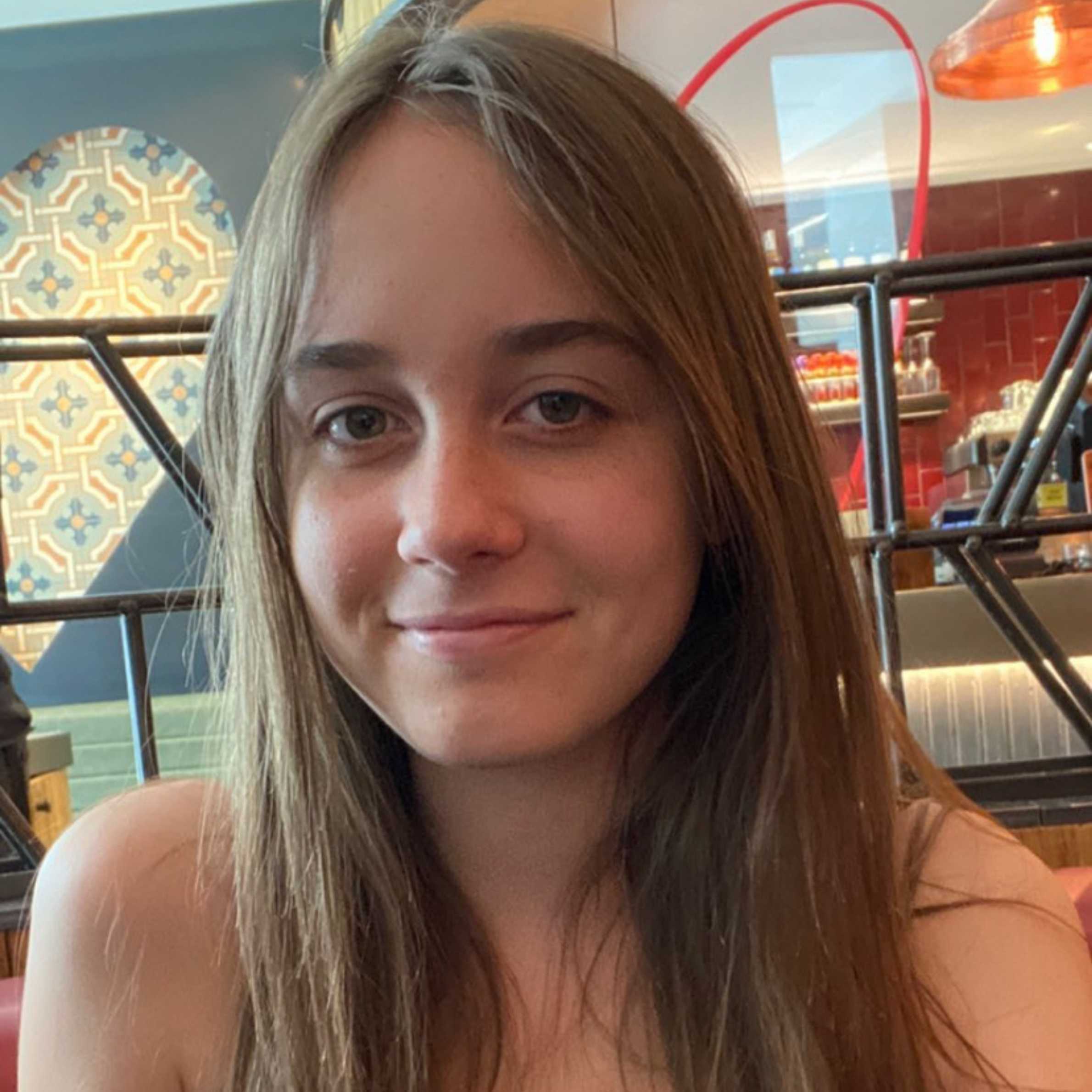 Girl with long light brown hair smiling at camera. She has brown eyes and eyebrows. In the background is a restaurant, colours of blue and red.