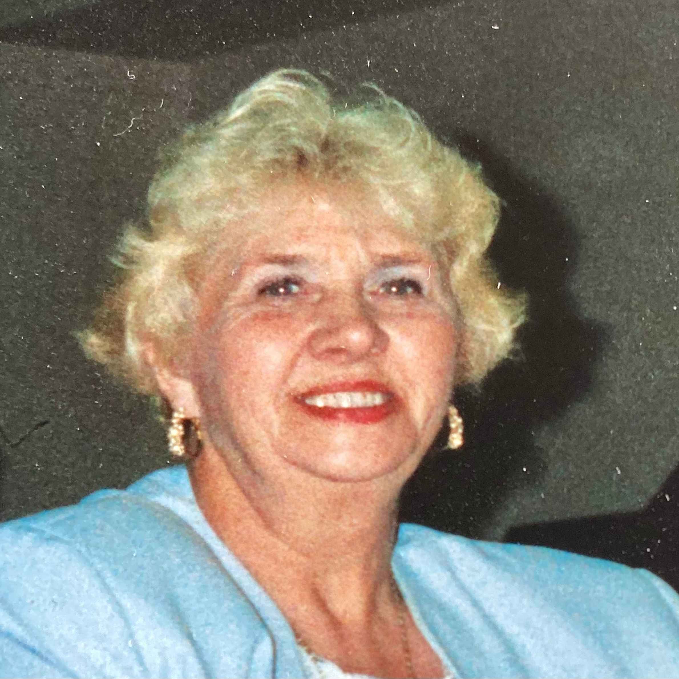 Lady smiling at camera, she is wearing red lipstick. She has blonde, short, curly hair and is wearing large chunky gold hoop earrings. She is wearing a blue jacket. The background is dark.