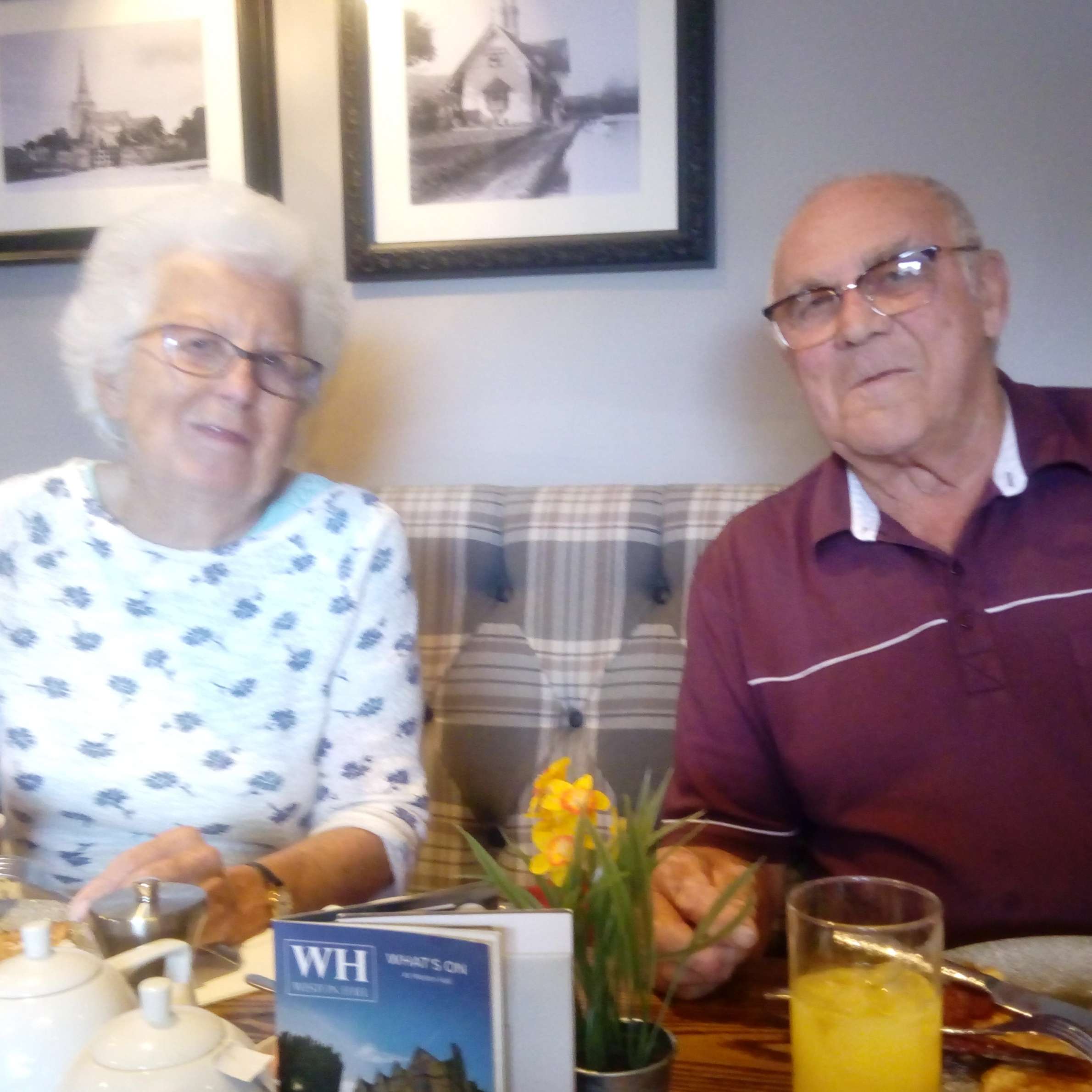 Lady sat to the left is wearing a white top with blue flowers on it, to the right is a man wearing a maroon coloured polo shirt. They are both wearing glasses and sat at a table in a restaurant eating.