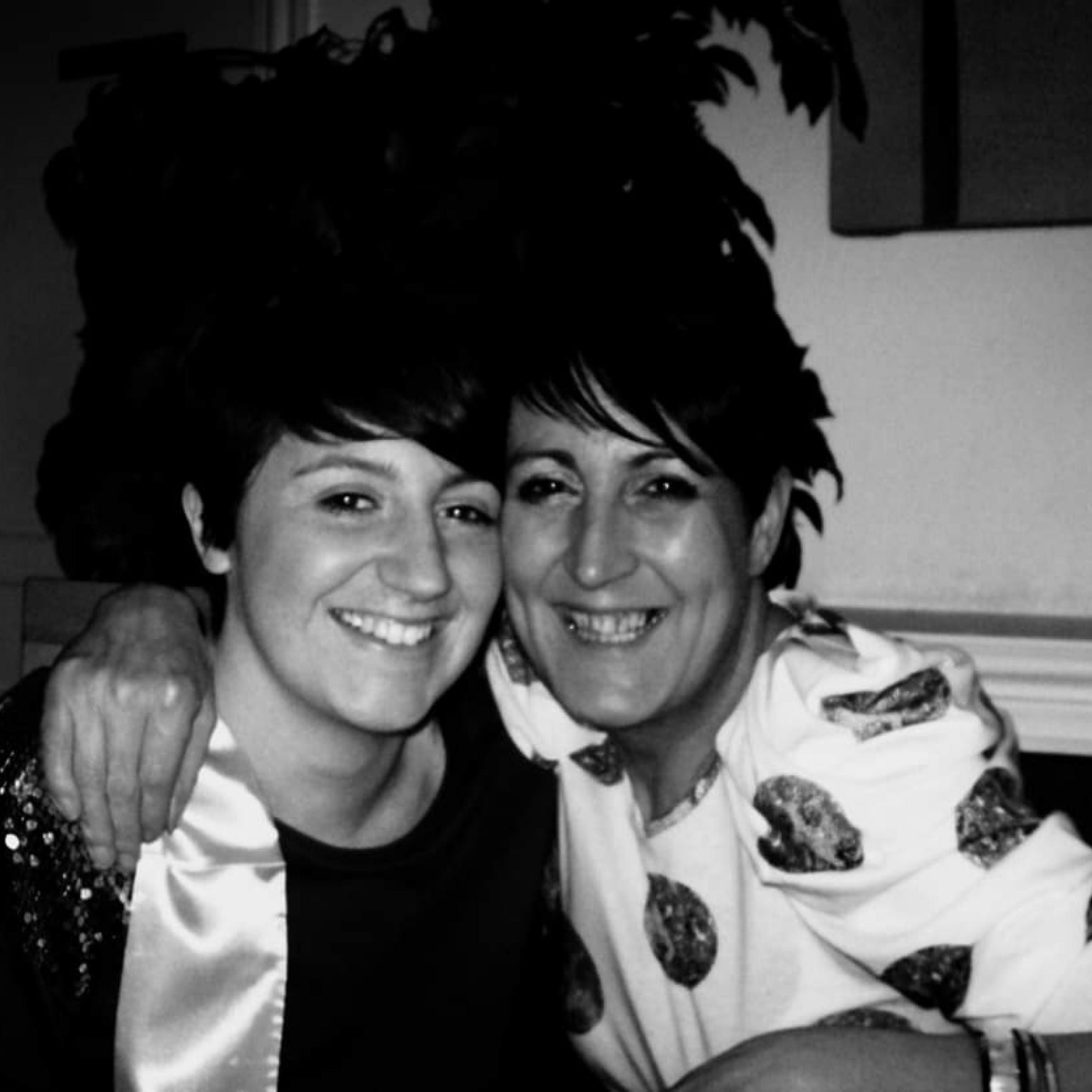 Black and white image of two ladies. They both have short dark hair, to the left the lady has a black top on with white sash. To the right the lady has a white and black spotted top on, she has her arm around the lady on the left.