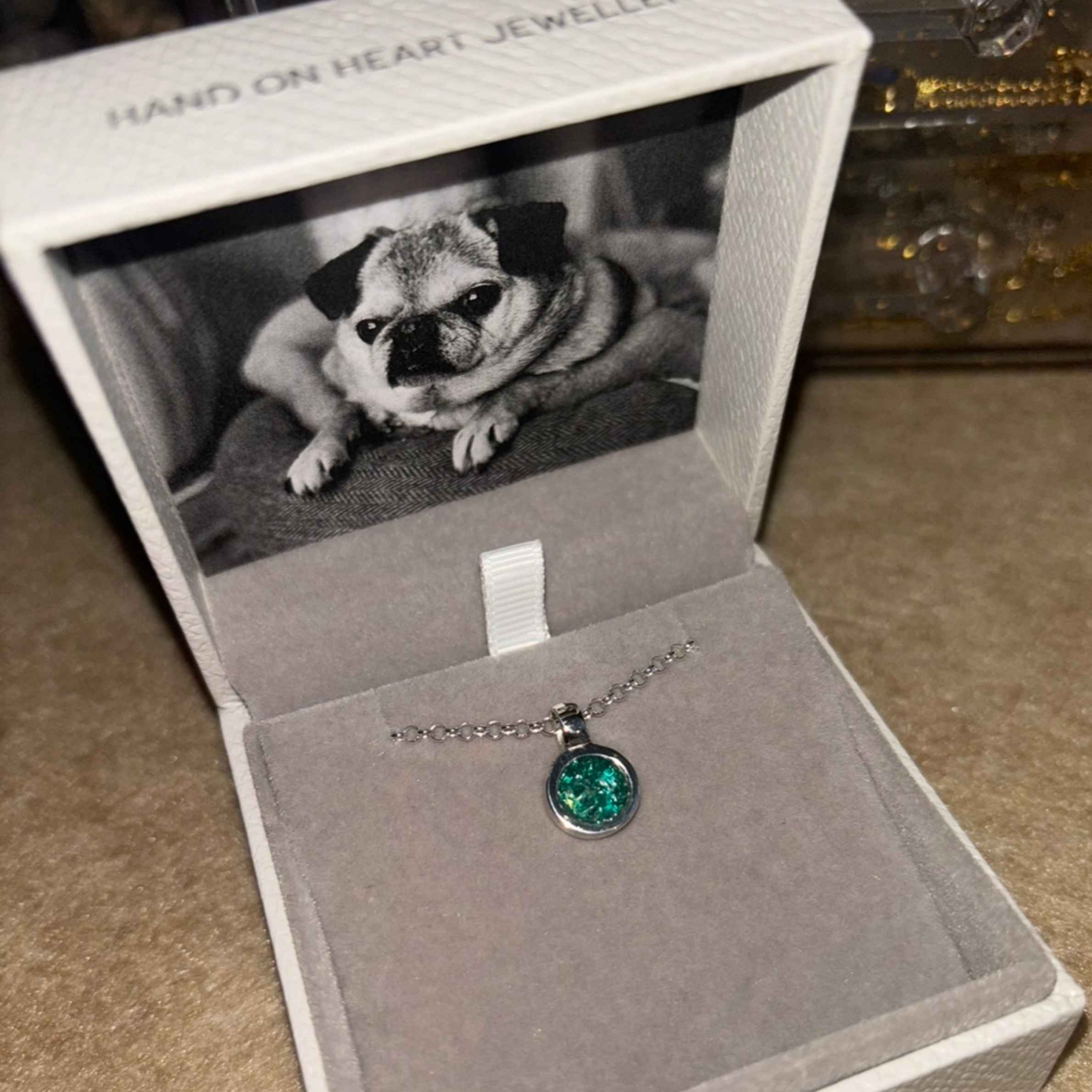 Photograph of green ashes necklace in Hand on Heart jewellery box. In the box there is a black and white image of a pug.
