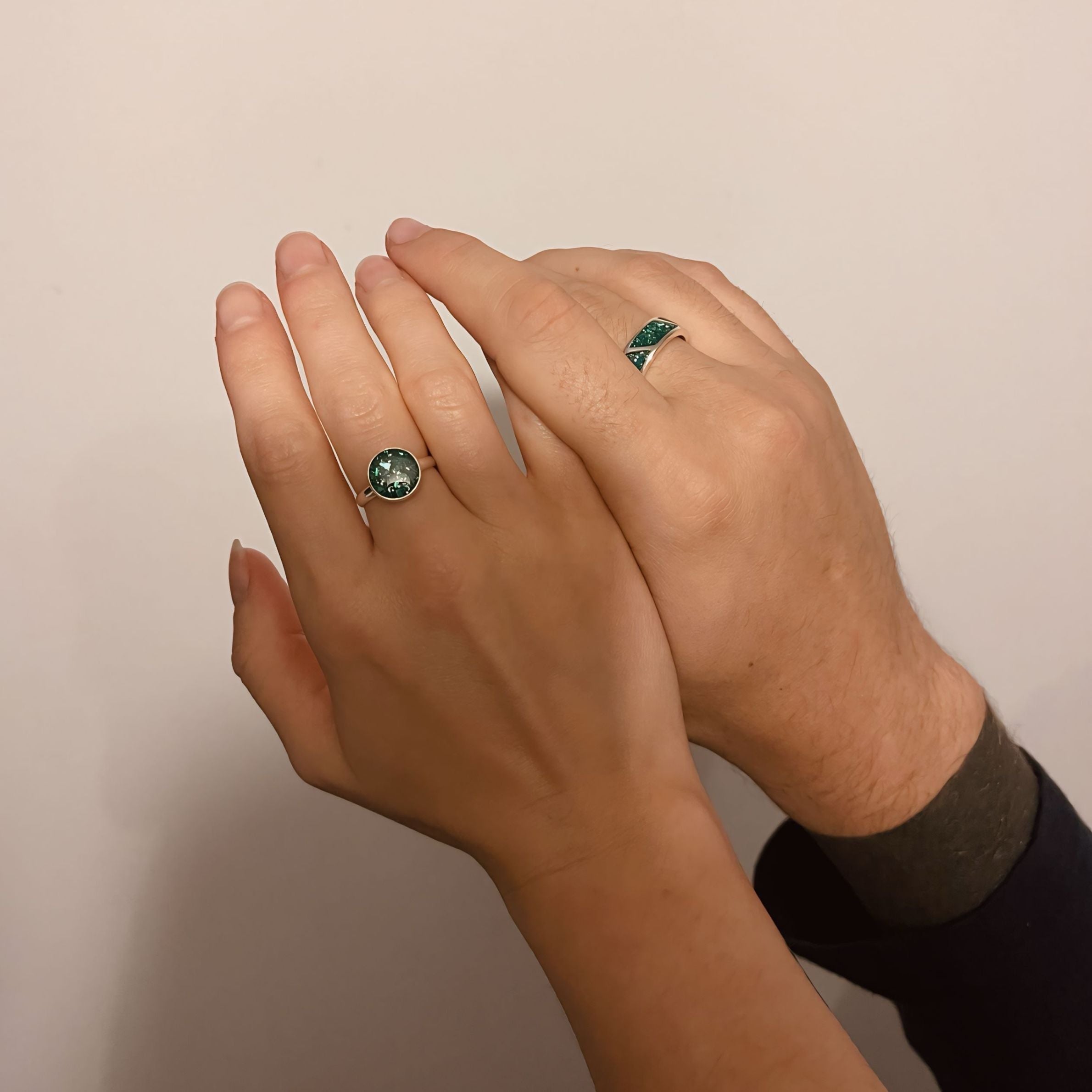 two hands touching one another against a white wall. one hand is a mans and the other a womans. Both wearing a green ashes rings