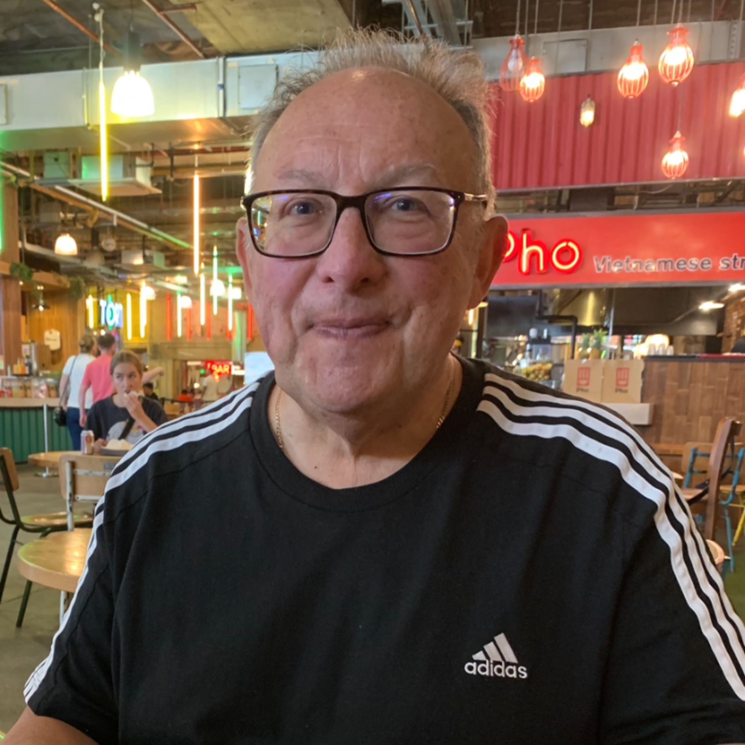 Man wearing a black t shirt with white striped sleeves and an adidas logo. hes wearing glasses and is in a restaurant setting with red and yellow lights