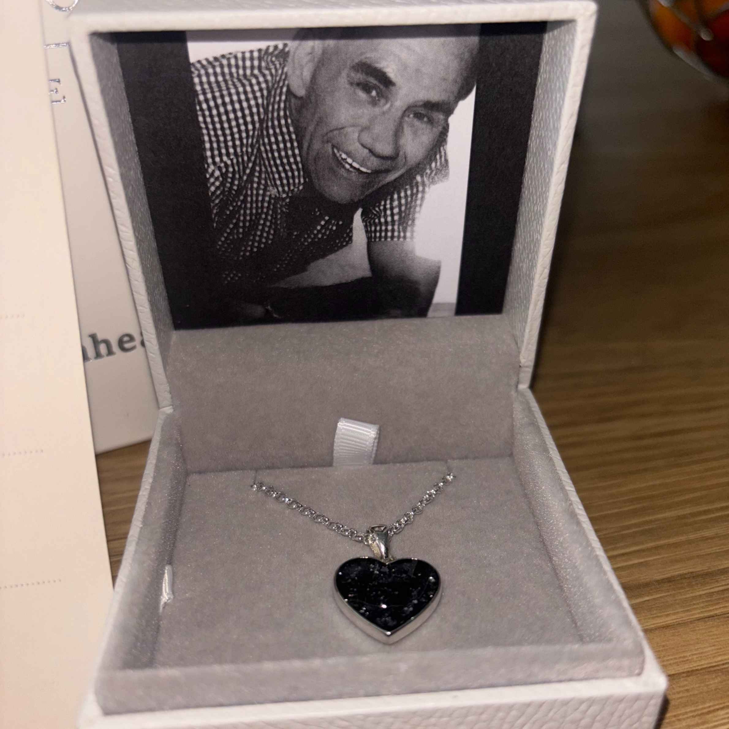 Photograph of black heart ashes necklace in Hand on Heart jewellery box. The jewellery box has a black and white image of a man smiling in a short sleeved checked shirt.
