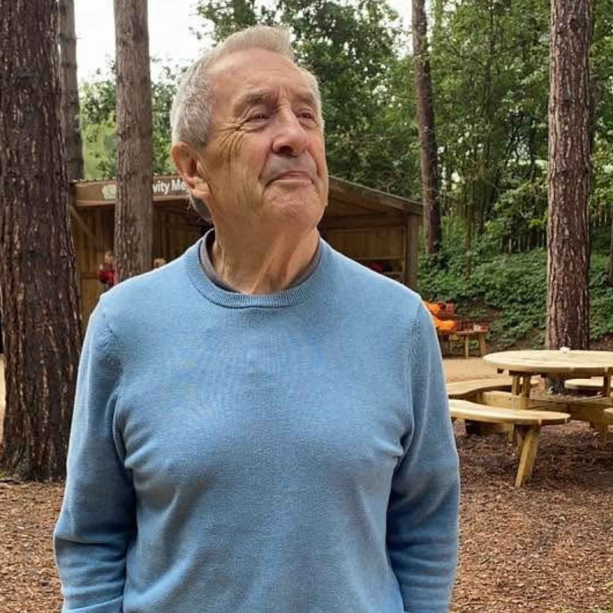 A man is stood in a forest looking ahead. He is wearing a blue crew neck jumper. There is a round picnic bench behind him.