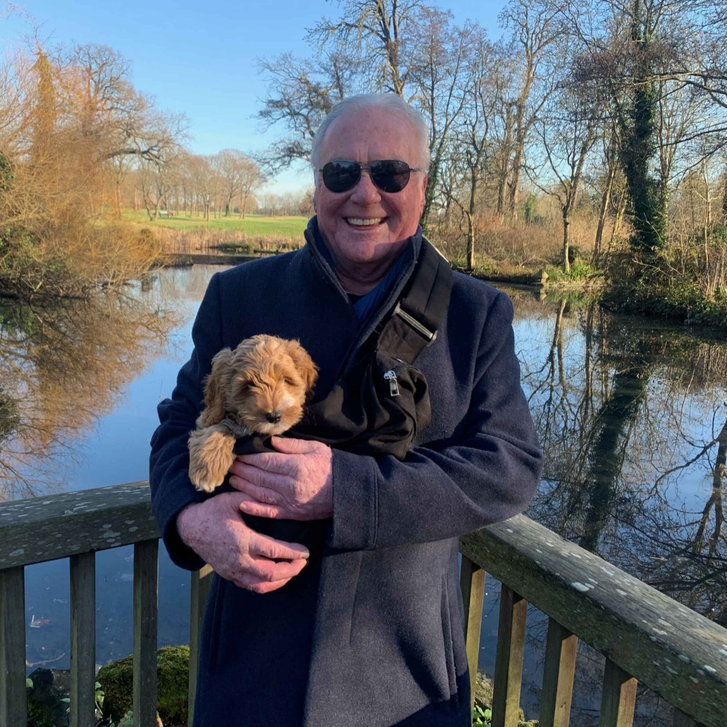 Man with white hair smiling. He is wearing a navy jacket and black sunglasses. He is holding a puppy. In the background is a lake and trees.