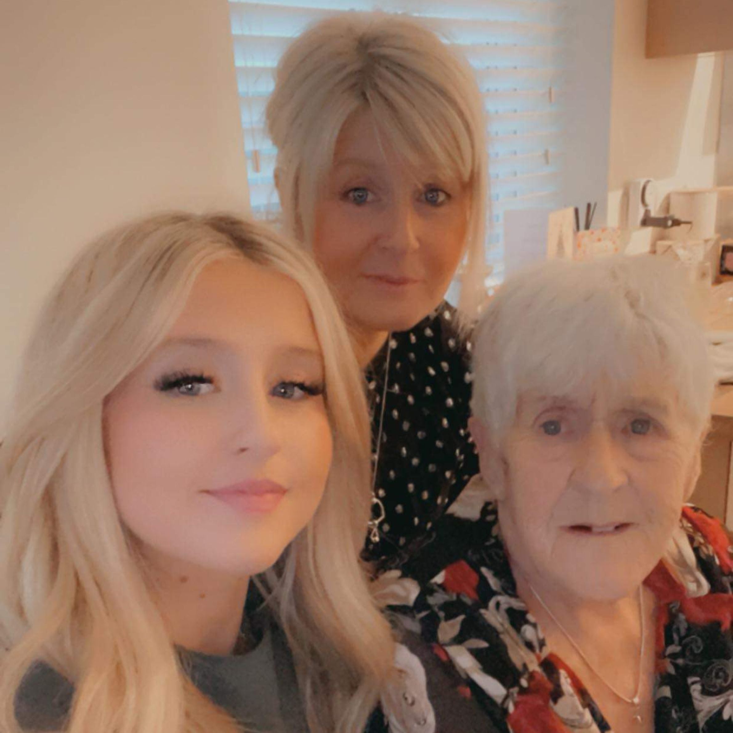 a selfie of three women from three generations. All with light hair and dark tops smiling for photo in front of cream wall with window.