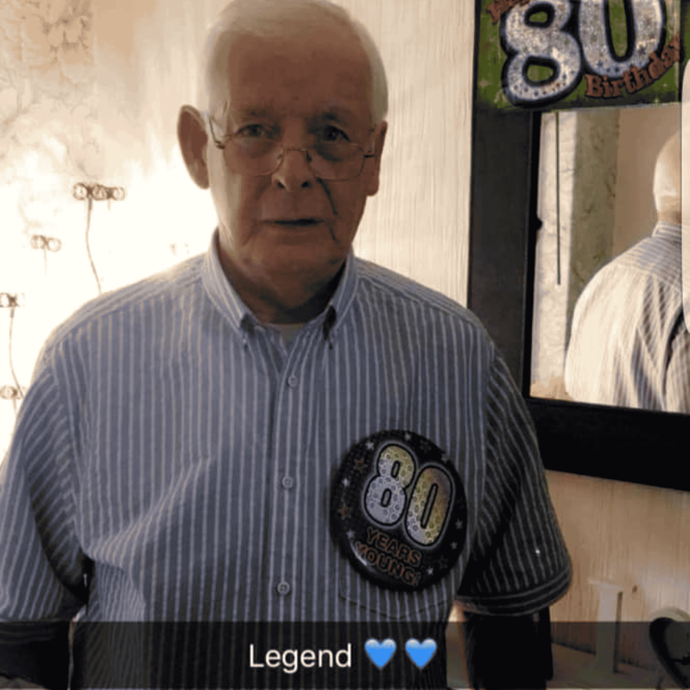 an older man with light grey hair wearing a blue and white striped shirt with a 80th birthday badge, there is a mirror in the background with an 80th birthday banner decoration, there is also text over the image saying "Legend 💙💙"