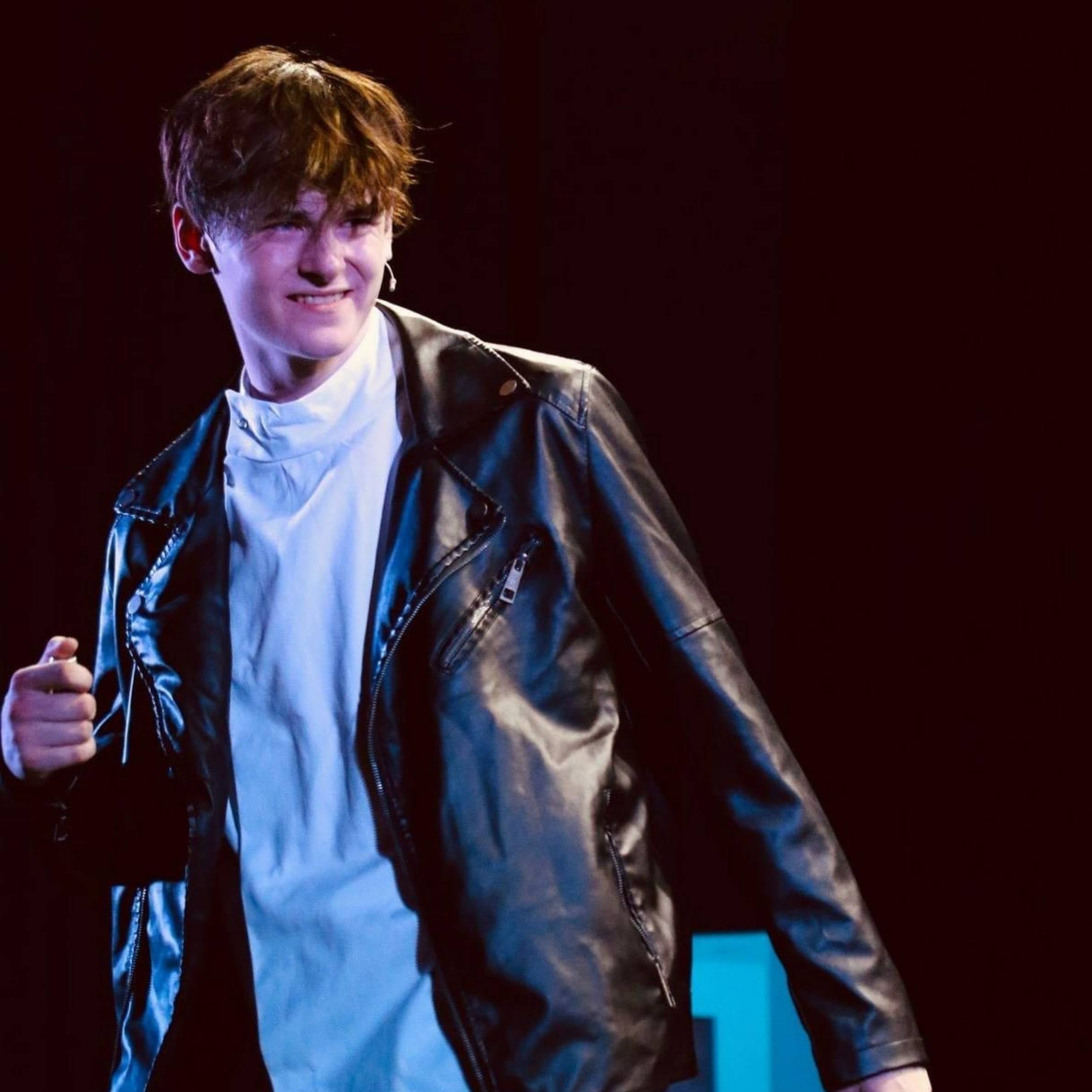 A young man wearing a white tshirt and black leather jacket is stood on a stage. His hair is brown and to his ears. The background of the image is dark.