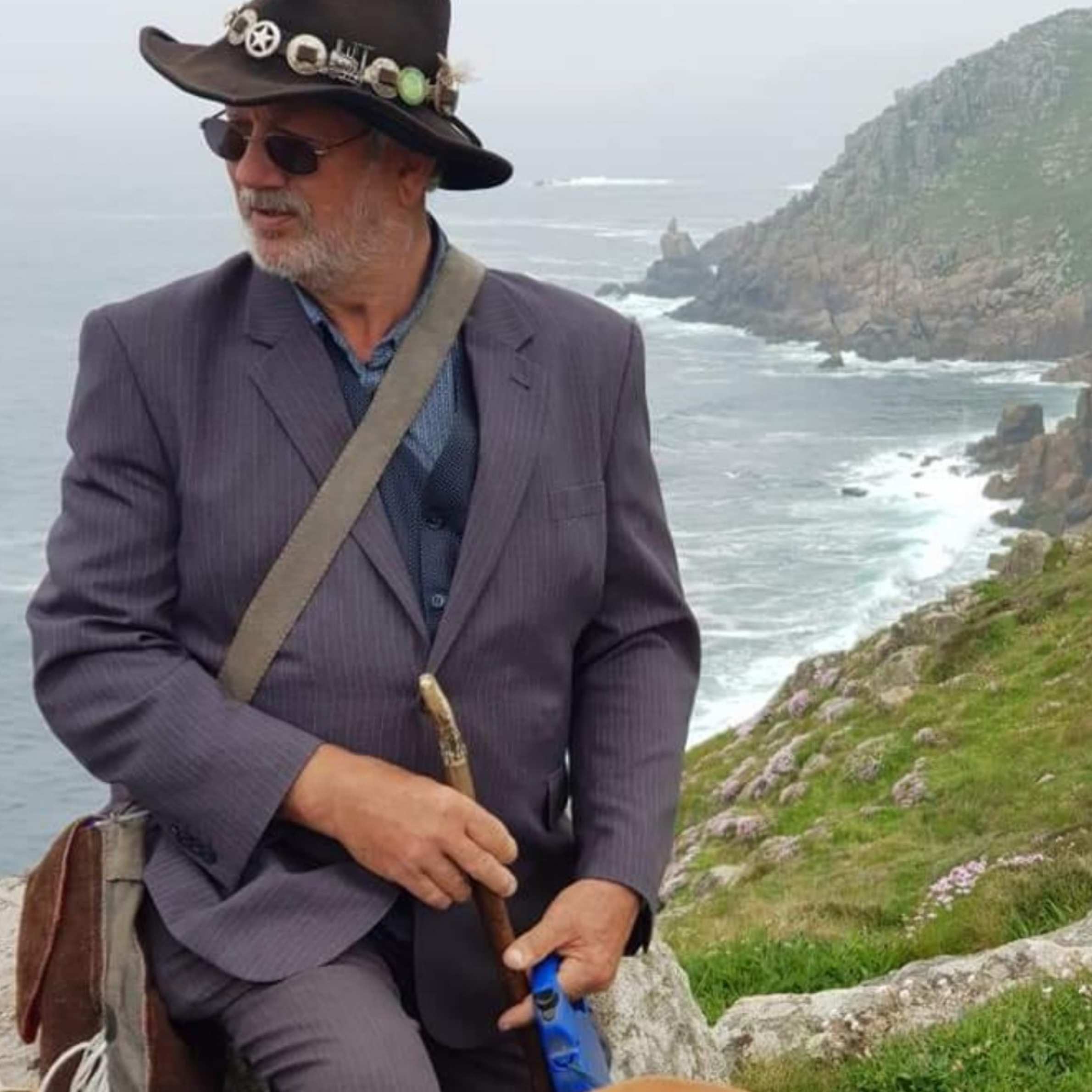 Man is wearing a navy blazer, cowboy hat and holding a wooden walking stick. He has a cross body bag across hsi front. In the background there is the sea and some cliffs.