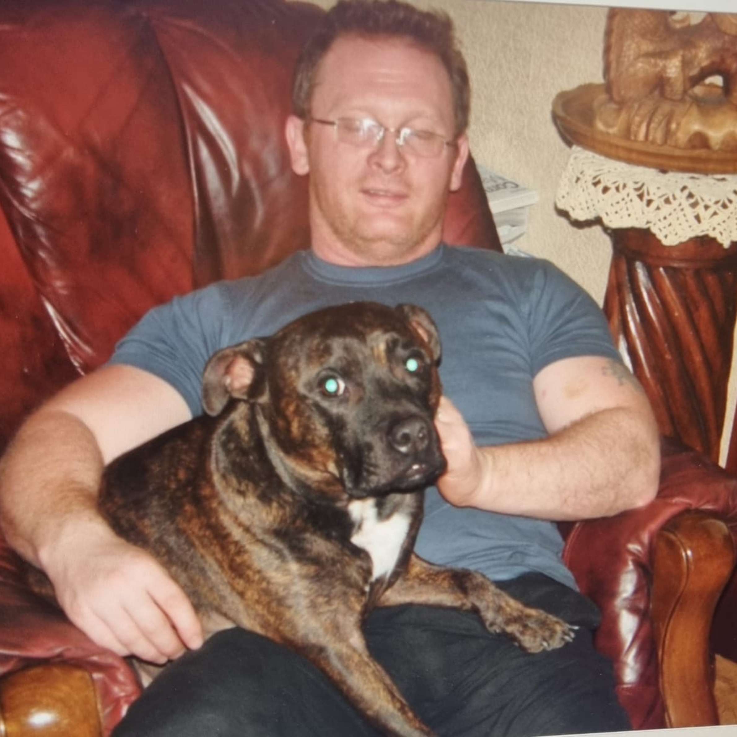 Man is sat with dog on his knee, on red leather chair. The man is wearing a short sleeved blue t-shirt and navy blue trousers. The dog is dark brown and black in colour with a white chest.