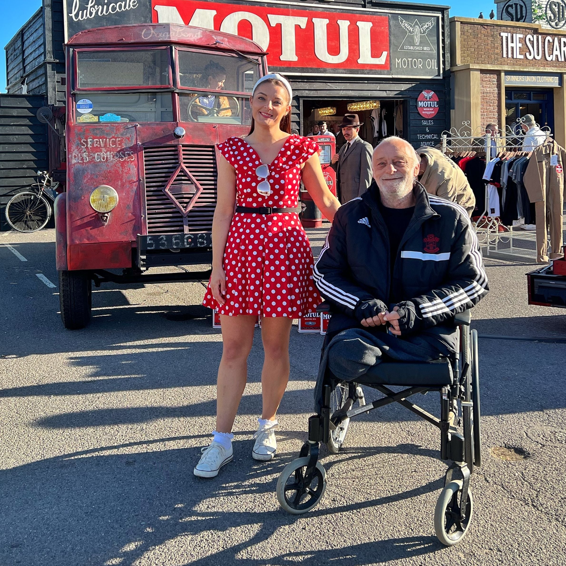 Girl in a red spotty dress standing next to a man wearing a black jacket in a wheelchair. There is a red truck in the background and a shop with a big red sign.