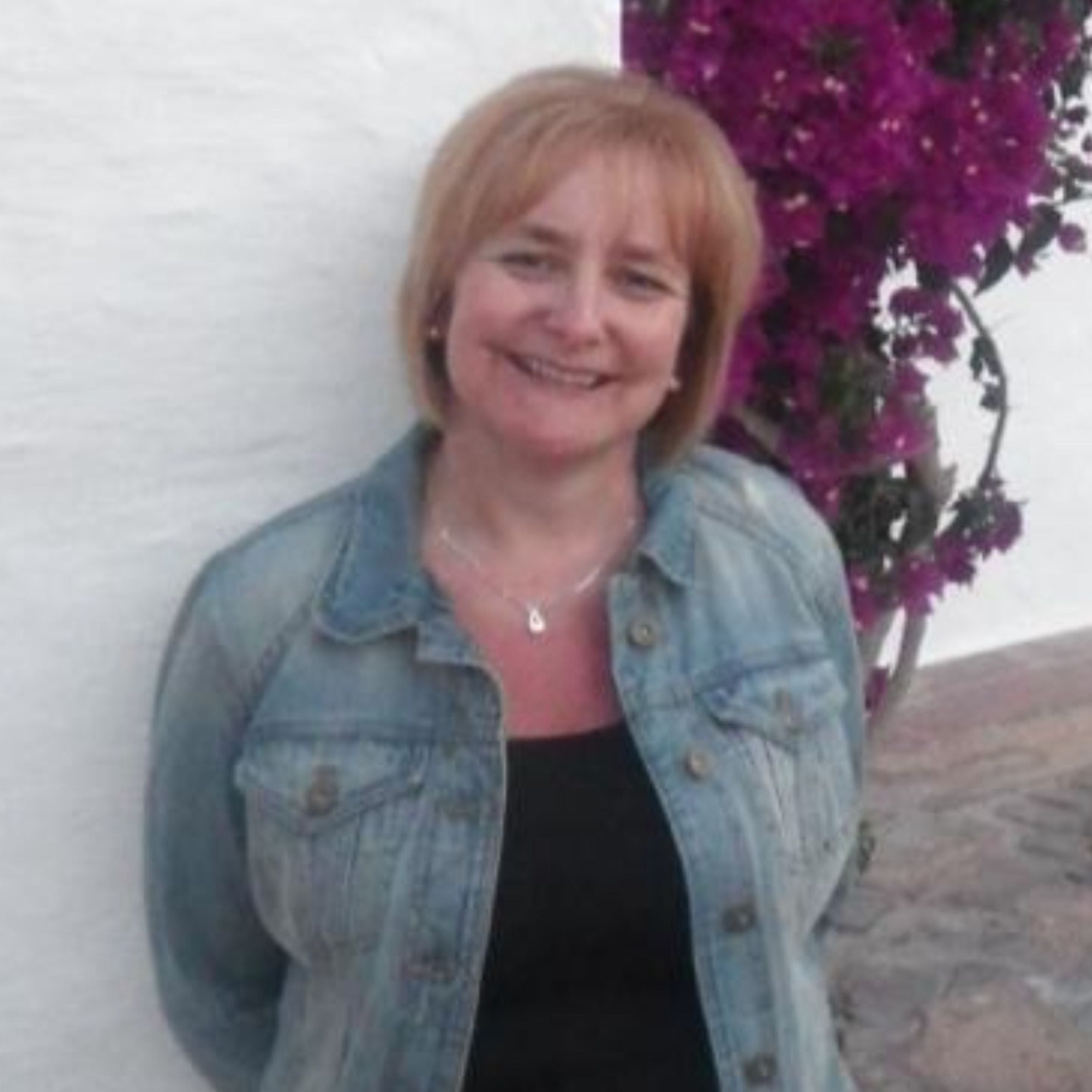 woman standing in front of a white wall and purple flowers. she is wearing a blue denim jacket and a black shirt. she is smiling and has reddish blonde hair