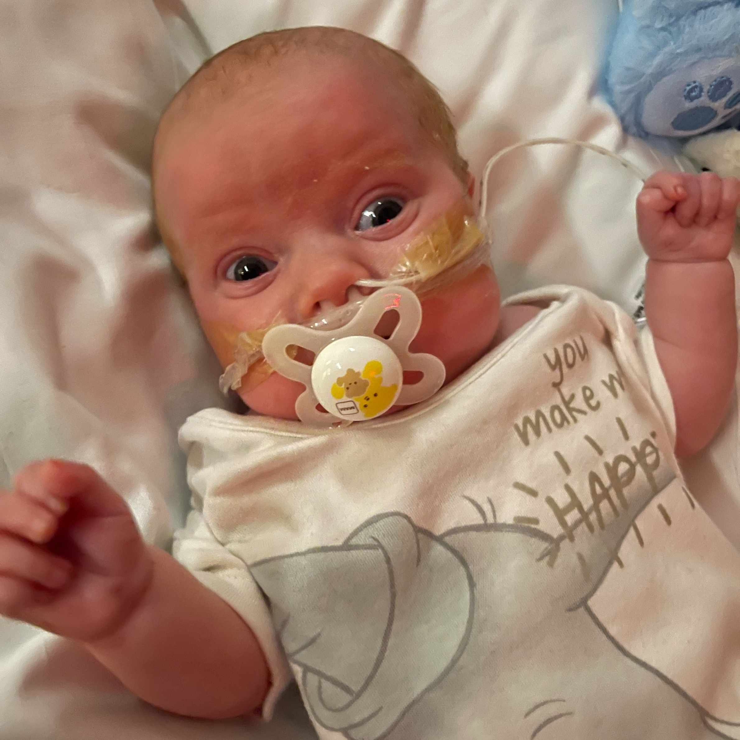 Baby laid on a white sheet, wearing baby grow with the words 'you make me happy' on the front. The baby had a yellow and white dummy in and is looking at the camera.