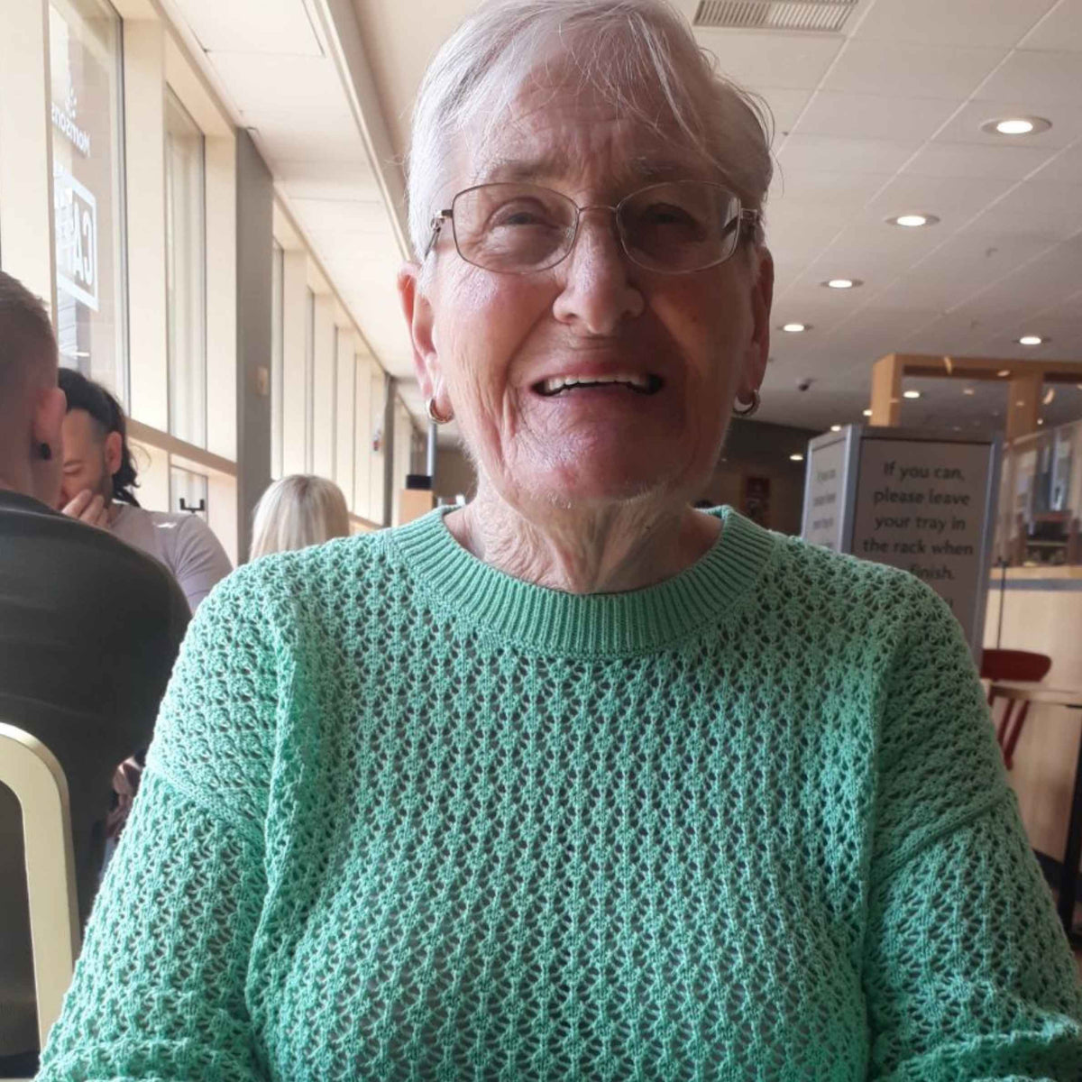 Smiling older woman with short gray hair and glasses, wearing a mint-green sweater in a bright, welcoming café or restaurant.
