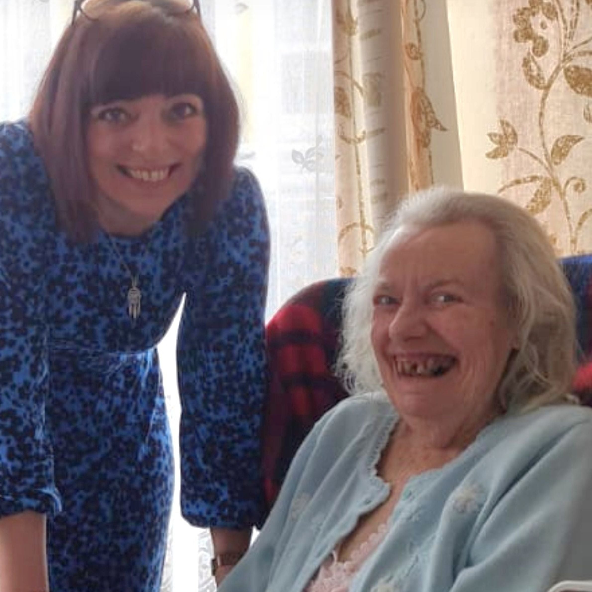 Two ladies, one with brown hair and purple top bending town for a photo with an older lady who is seated with a purple cardigan on and long grey hair
