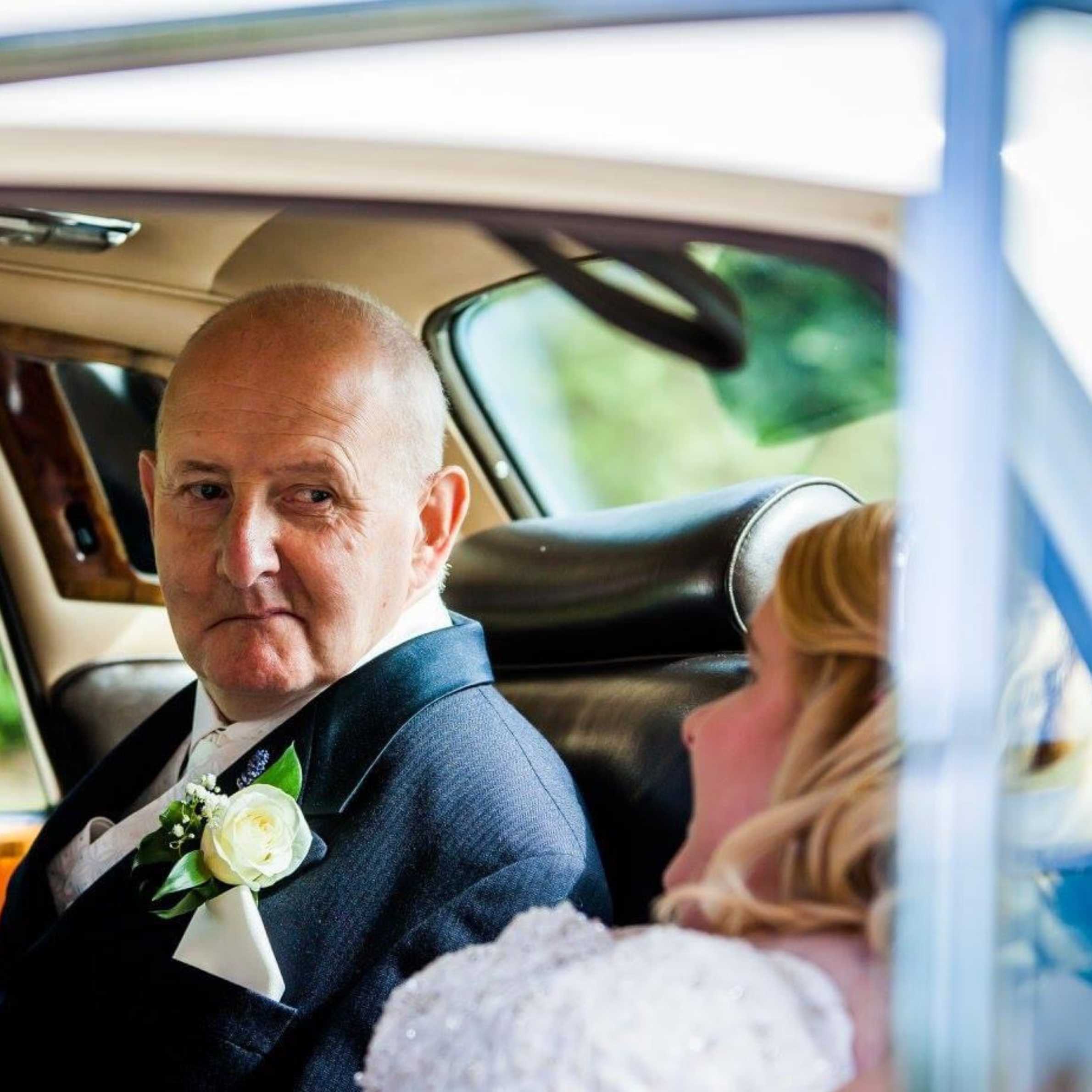 Bald man sat in the back of a car. He is wearing a navy suit, with a white rose buttonhole. There is a lady to the right of the photo with her back to the camera, wearing a white wedding dress with blonde hair.