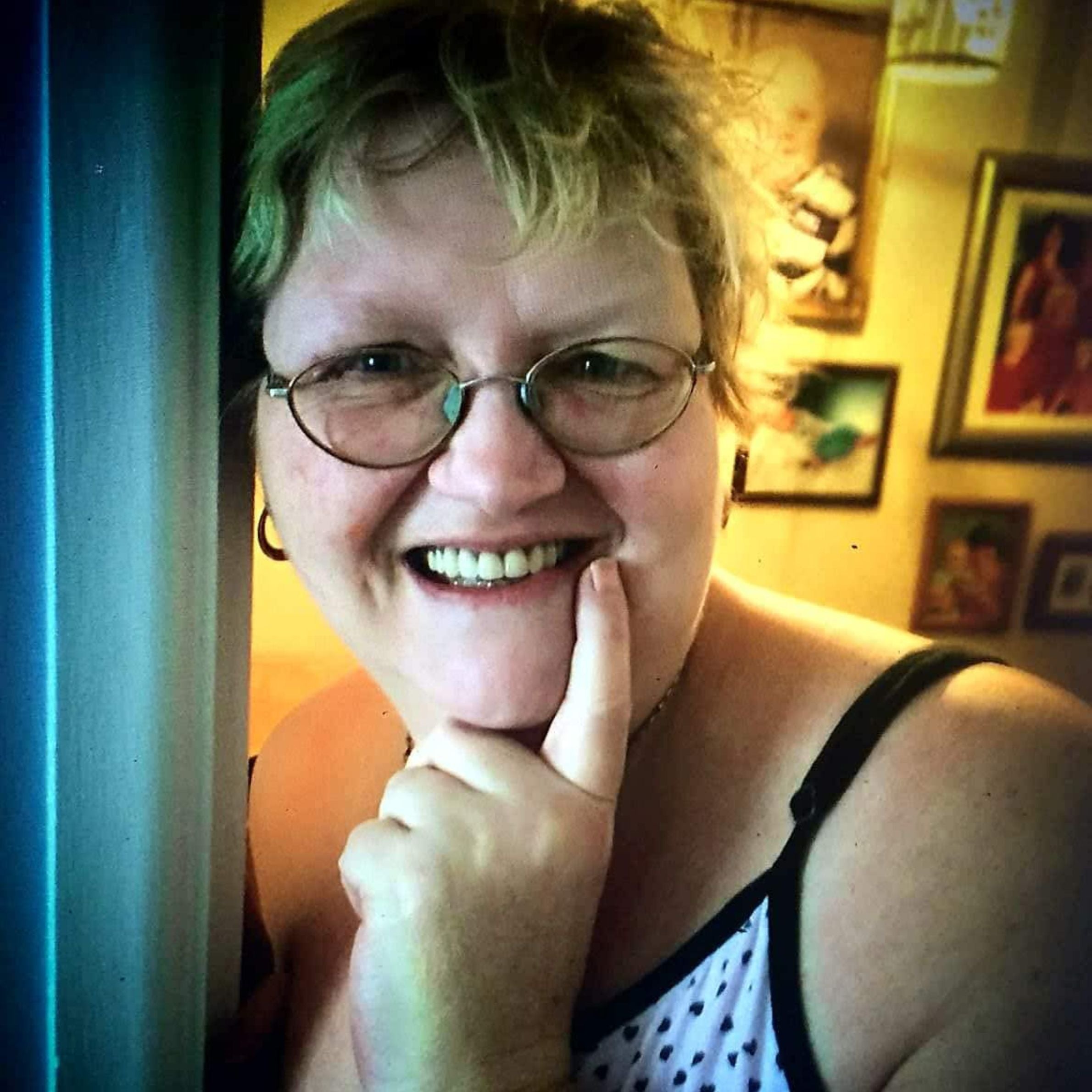 woman with short blonde hair smiling wearing glasses and a black and white dotted top. she is leaning on a door frame and there is a yellow wall with picture frames in the background