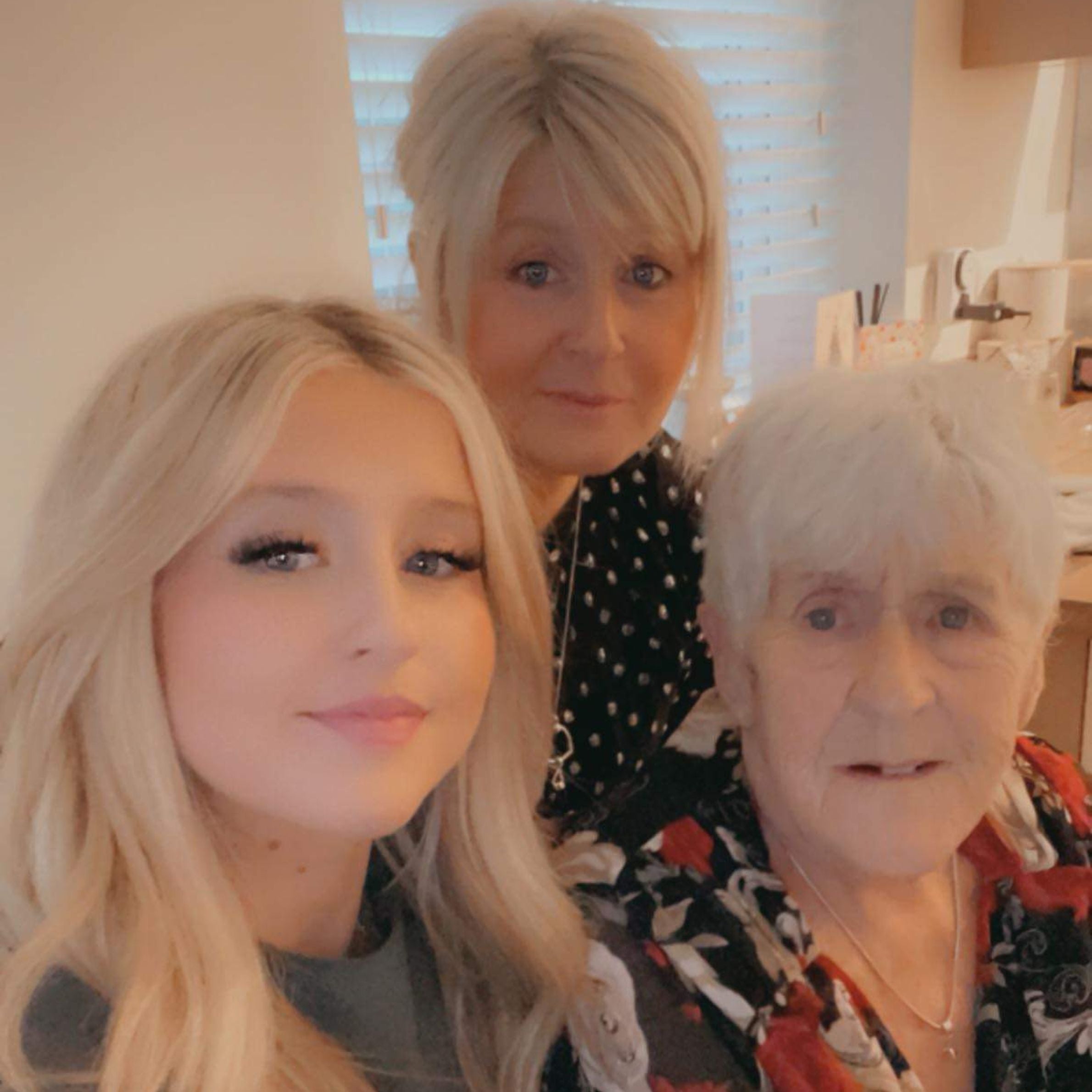 a young blonde girl wearing a grey jumper, a middle aged blonde woman wearing a black and white dotted top and an older woman with grey hair smiling for a photo. There is white window blinds in the background