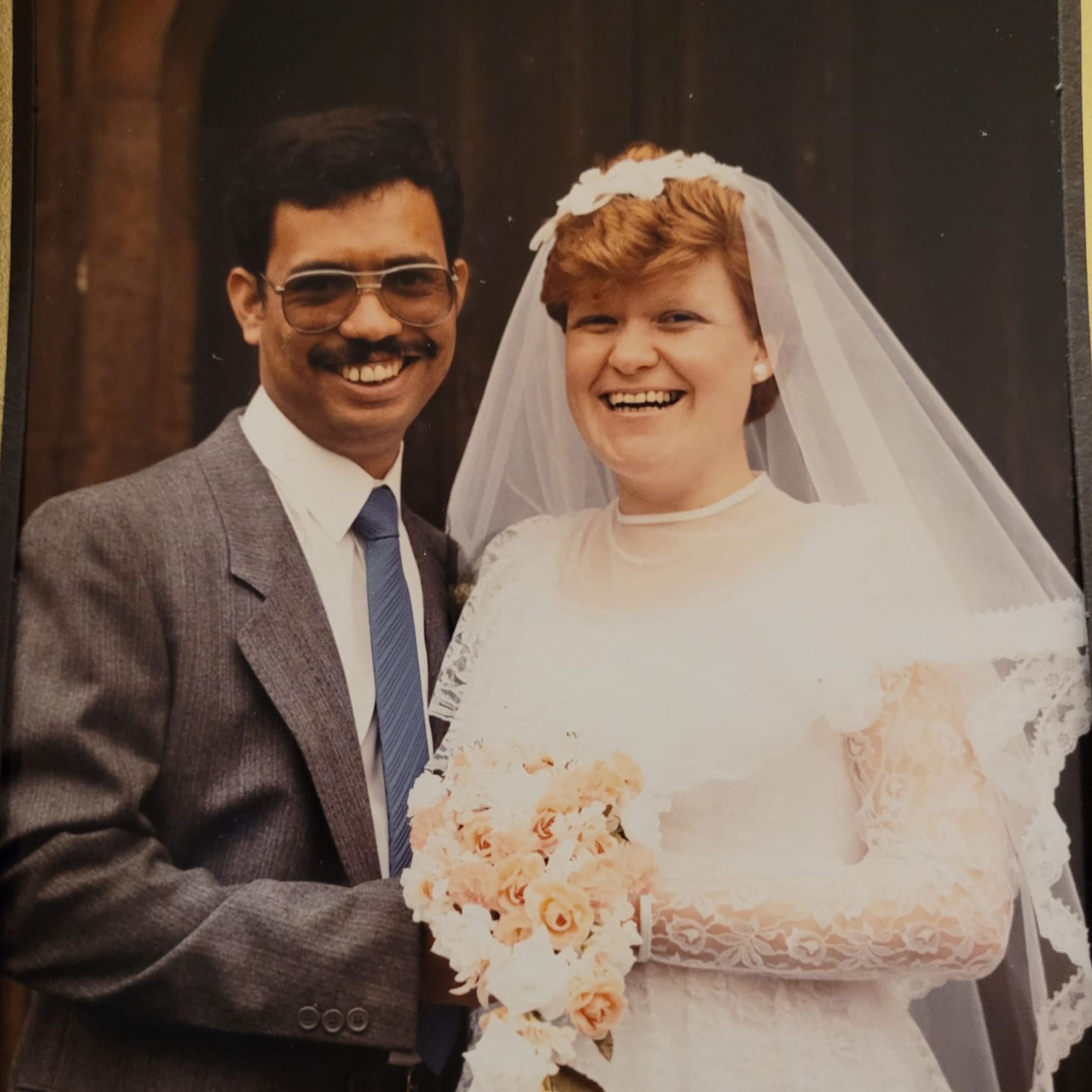 A bride and groom on their wedding day. The man is wearing a grey suit, blue tie and sunglasses and the woman is wearing a white wedding dress with a veil and a bouquet of white and peach flowers. The bride has red hair.