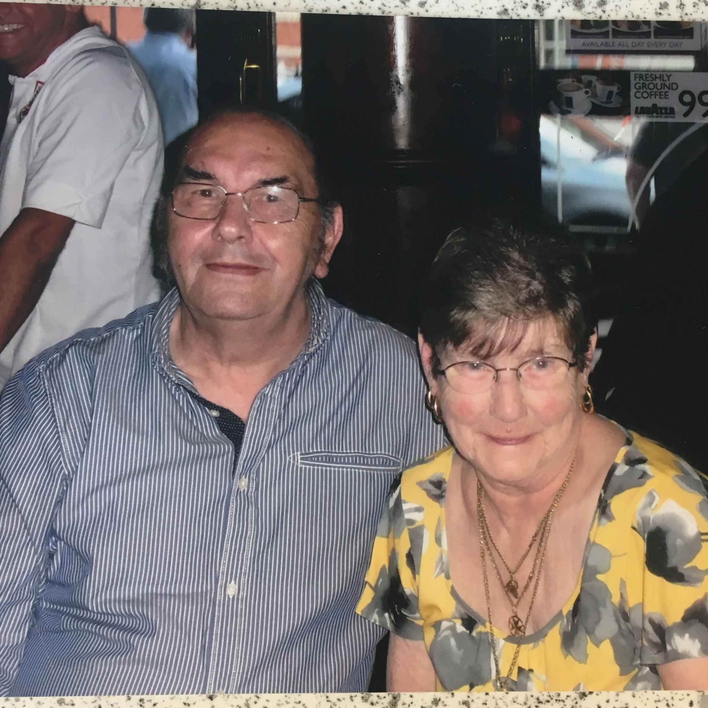 Man in blue striped shirt sat next to lady in grey and yellow floral blouse. They are both wearing glasses and smiling. Dark background with distant man in white shirt.