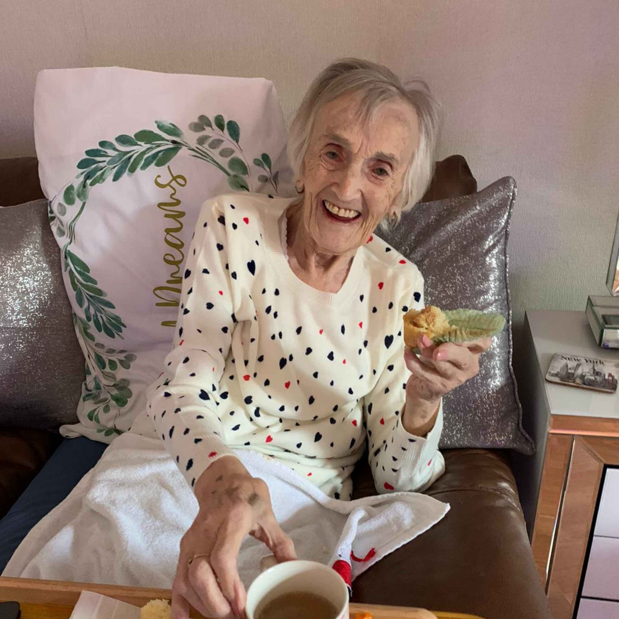 Lady smiling at camera, she is sat on a brown leather seat, holding cupcake and mug. She is wearing a cream jumper with multicoloured spots on it. There is a white pillow behind her with green leaves and gold writing on.