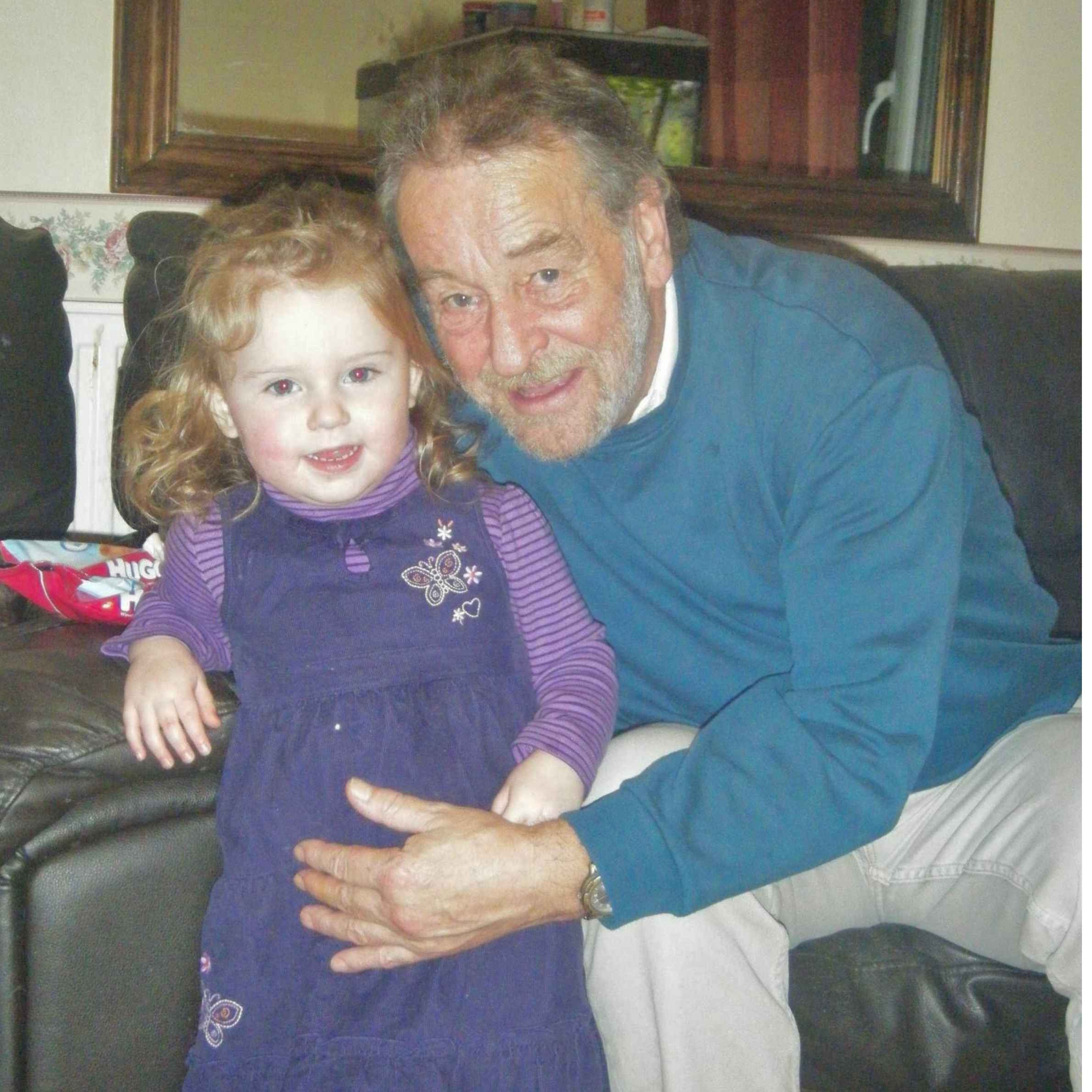 Older man wearing a blue jumper and grey trousers holding a little girl wearing a purple dress and ginger hair. they are sat on a dark sofa and there is a mirror on the wall in the background