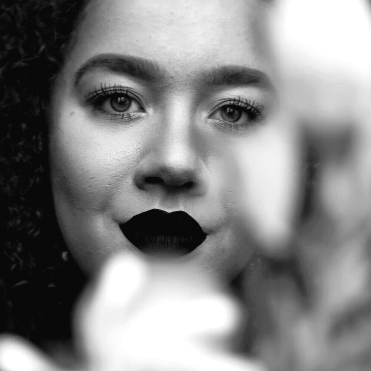 A black and white image of a young lady facing the camera. She is wearing dark lipstick and has large full eyelashes. She is facing towards the camer while an object is out of focus in the foreground