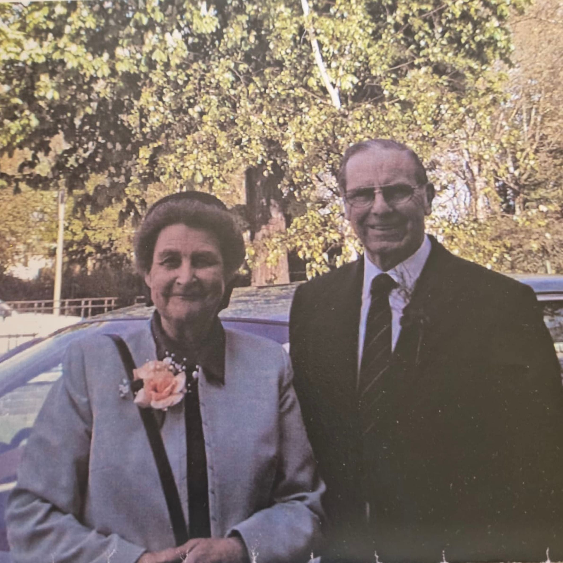 an older woman with short hair wearing a grey coat with a pink flower standing next to an older man wearing a black suit with a striped tie wearing glasses. there is trees in the background