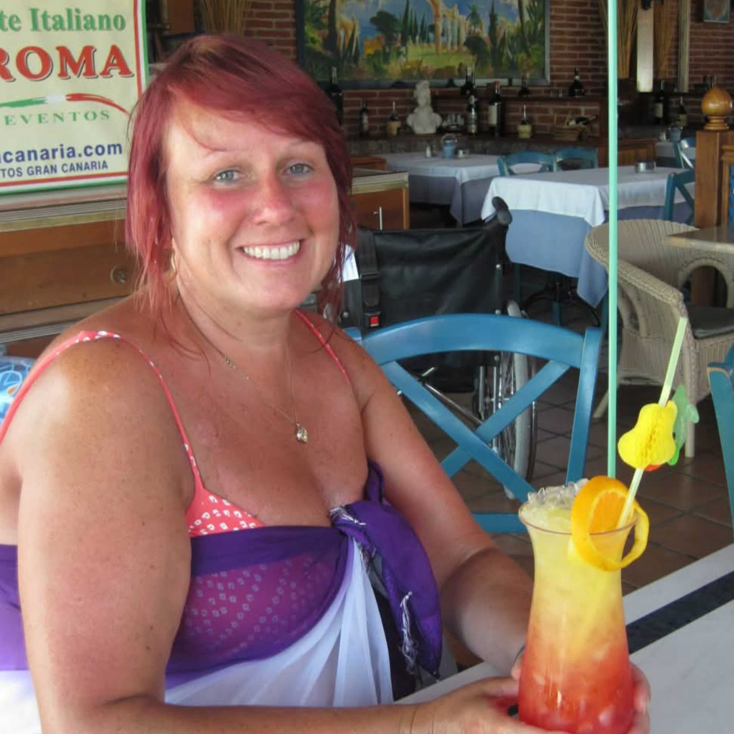A woman is sat at table with a yellow and red cocktail with a bright colured straw. She has short red hair and is wearing a pink bikini top with a purple top over it.