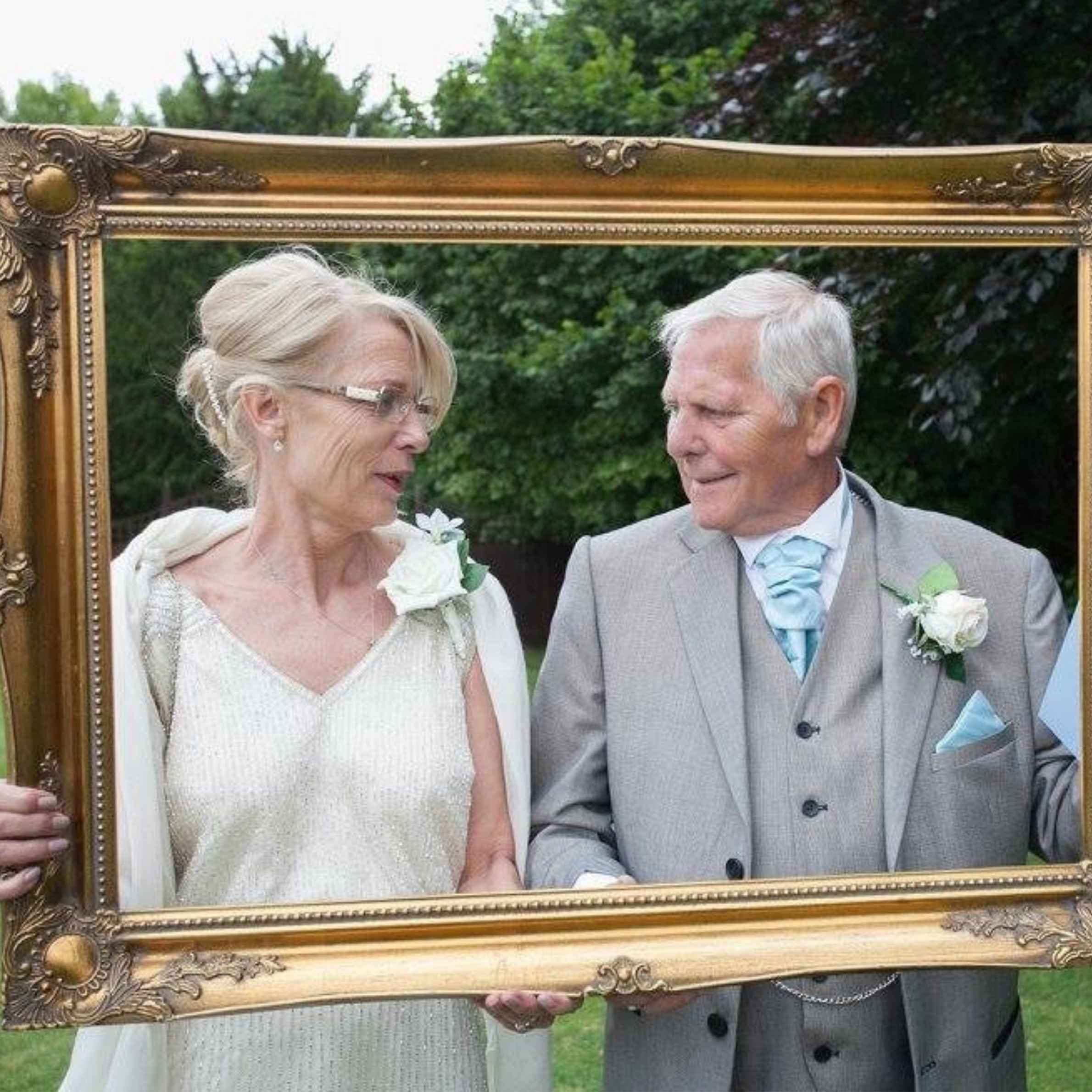 Lady in cream sparkly dress to the left of the photo, to the right a man in a grey suit with blue tie. They are holding a large gold photo frame. Trees in the background.