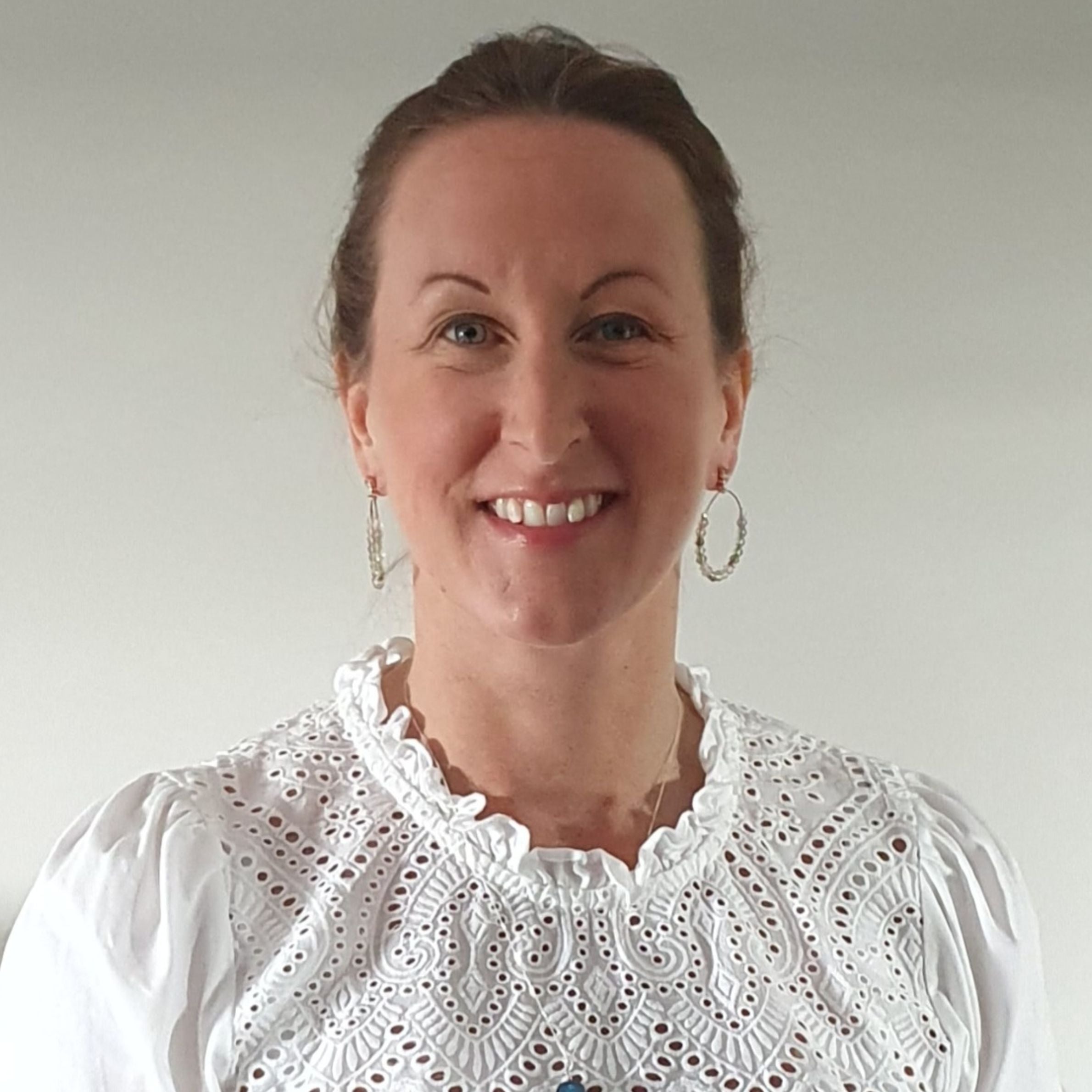 woman smiling for a photograph wearing a white lace shirt and hooped earrings. she has light brown hair stood against a blank pale background