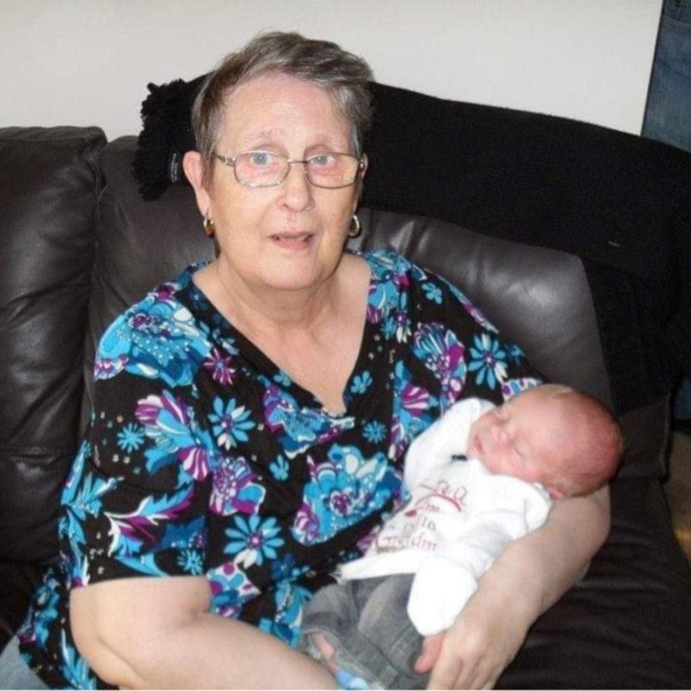 An older woman wearing glasses with short grey hair wearing a black shirt with blue and purple flowers on it. She's holding a baby wearing a white top while sitting on a dark coloured sofa