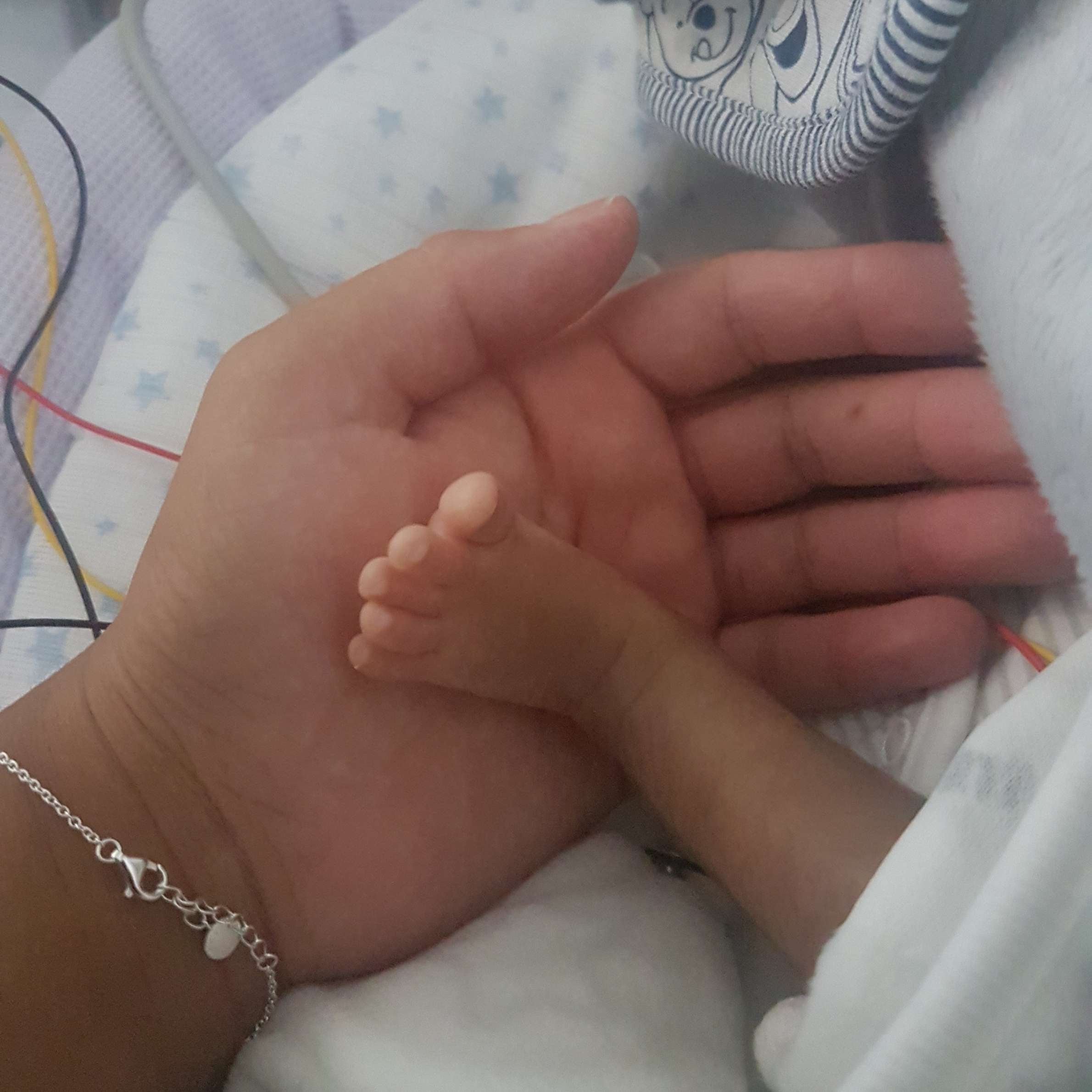 Picture of baby's foot held by hand. Hand has silver bracelet on wrist. The background is light in colour, a baby blanket.