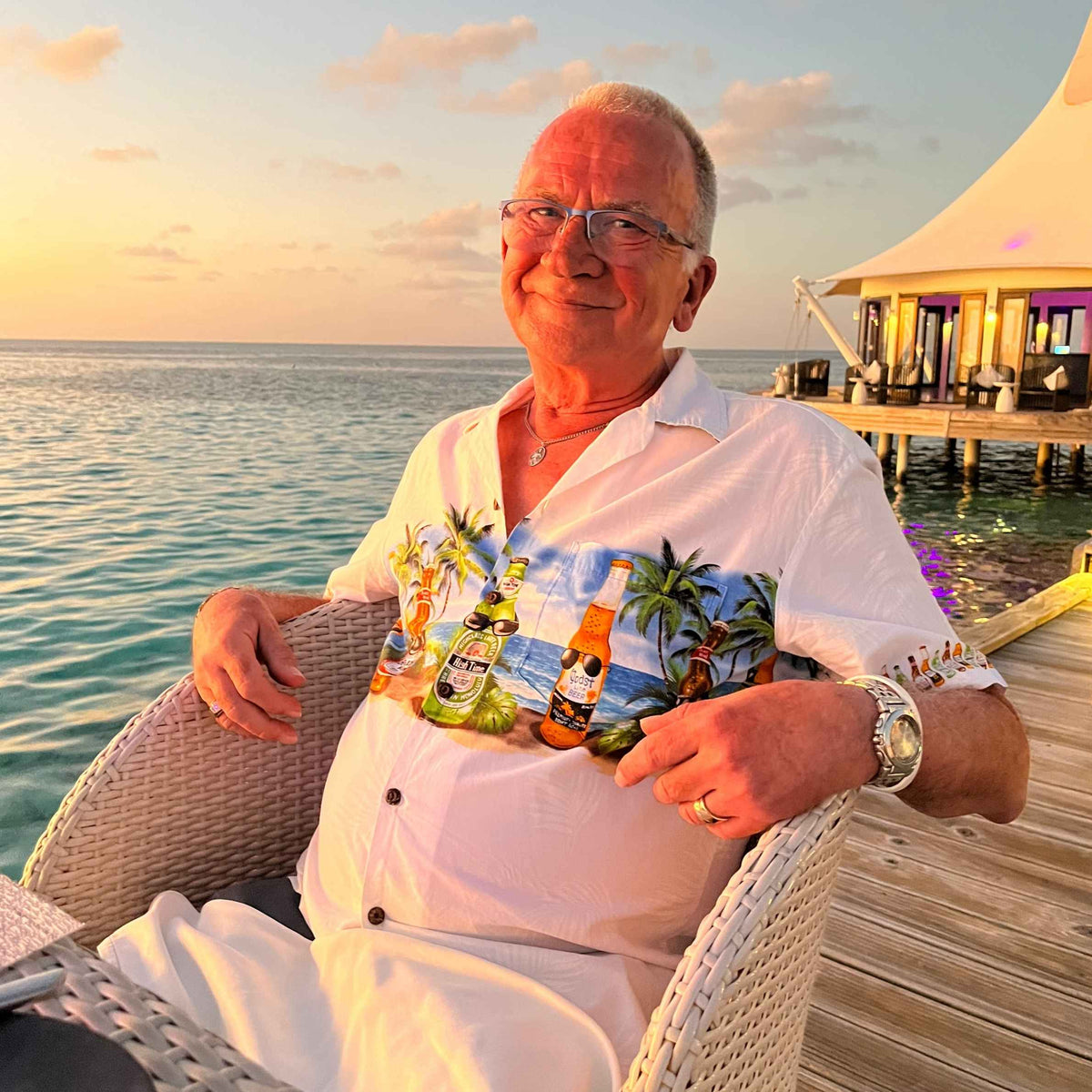 Smiling older man in a colorful floral shirt relaxes in a wicker chair with drinks at a tropical seaside resort during sunset.