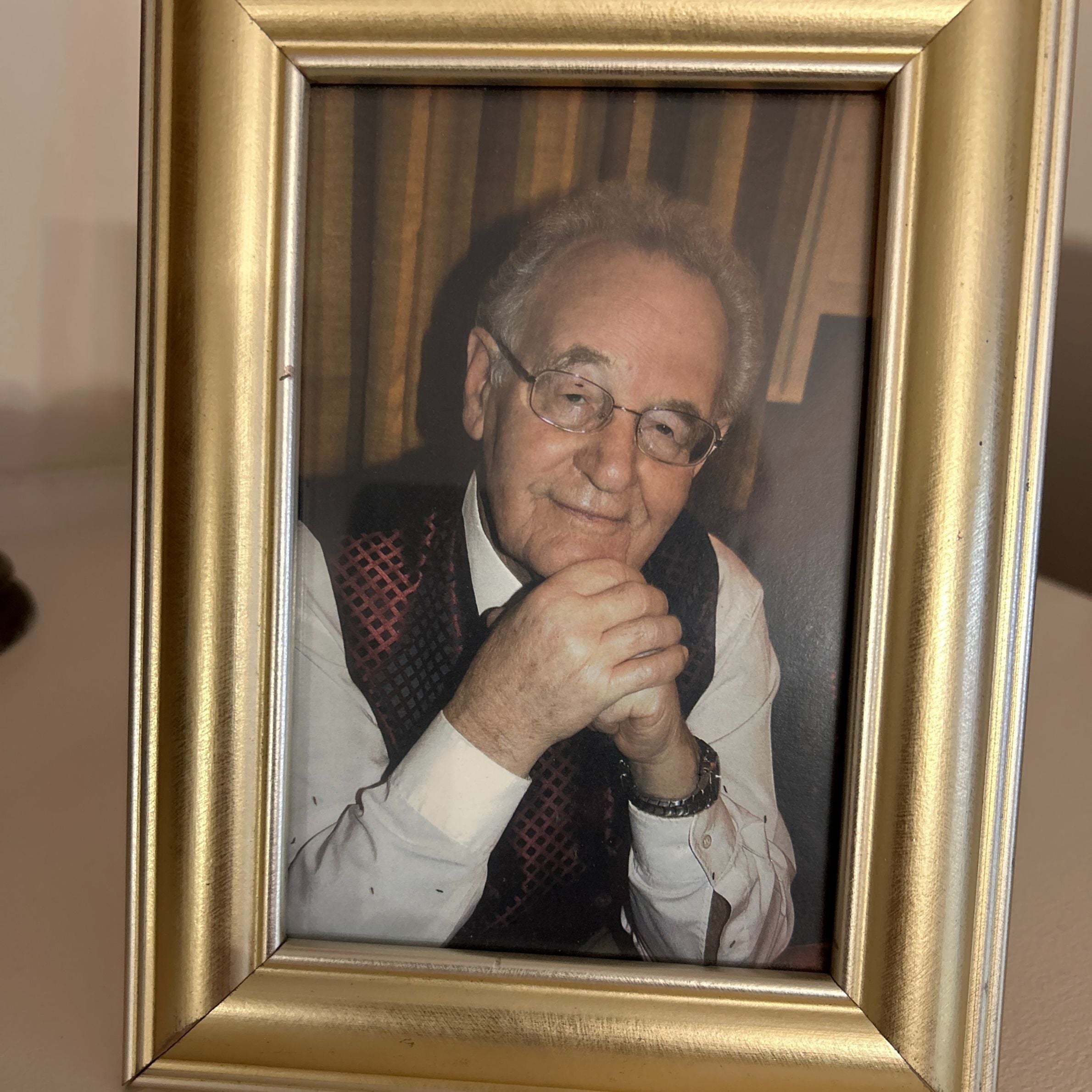 gold photo frame holding a picture of an older man wearing a white shirt and a dark red waist coat. he has grey hair and is wearing glasses and smiling