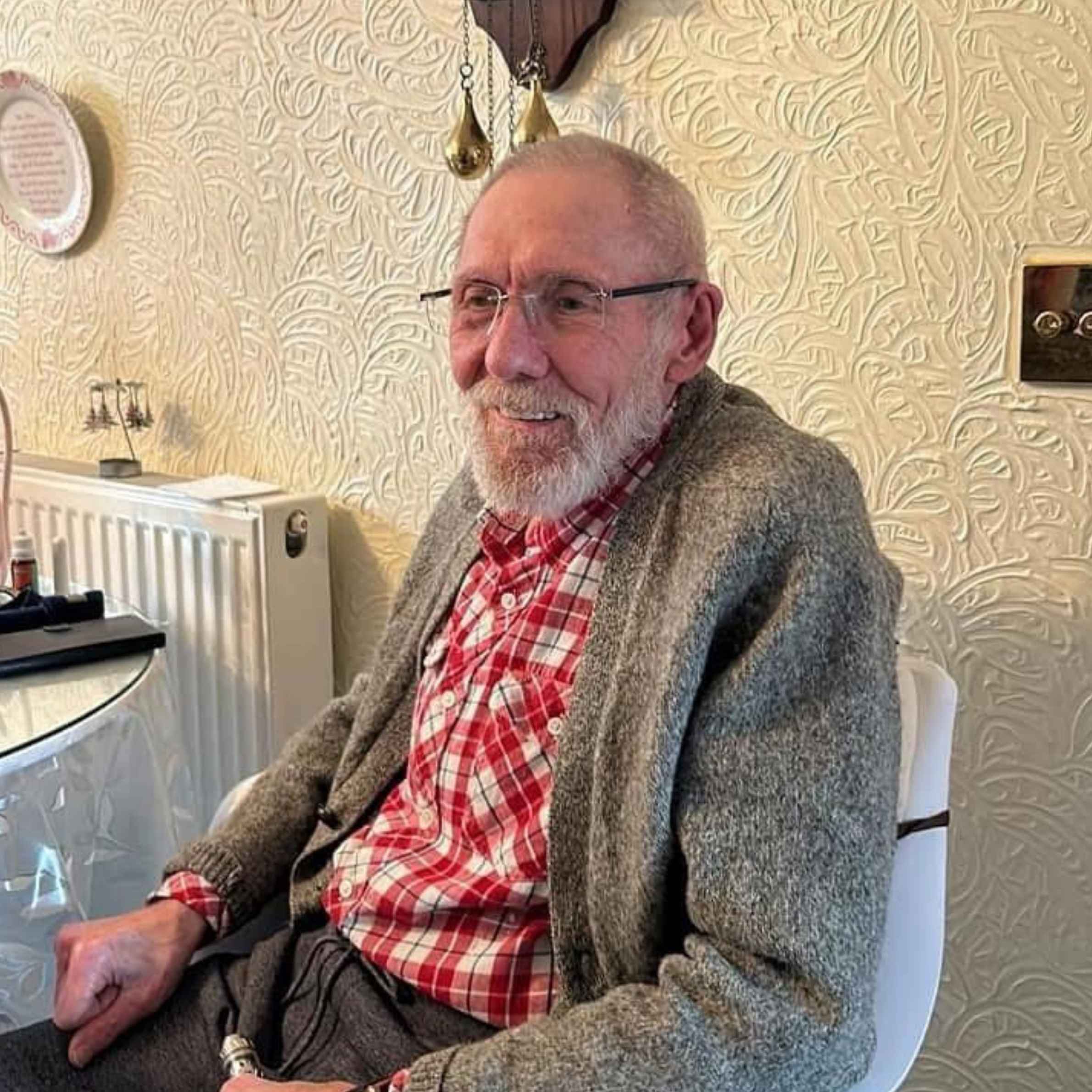 An older gentleman sat in a white chair at a table next to the radiator. The man is wearing glasses and a red checked shirt with a grey cardigan.