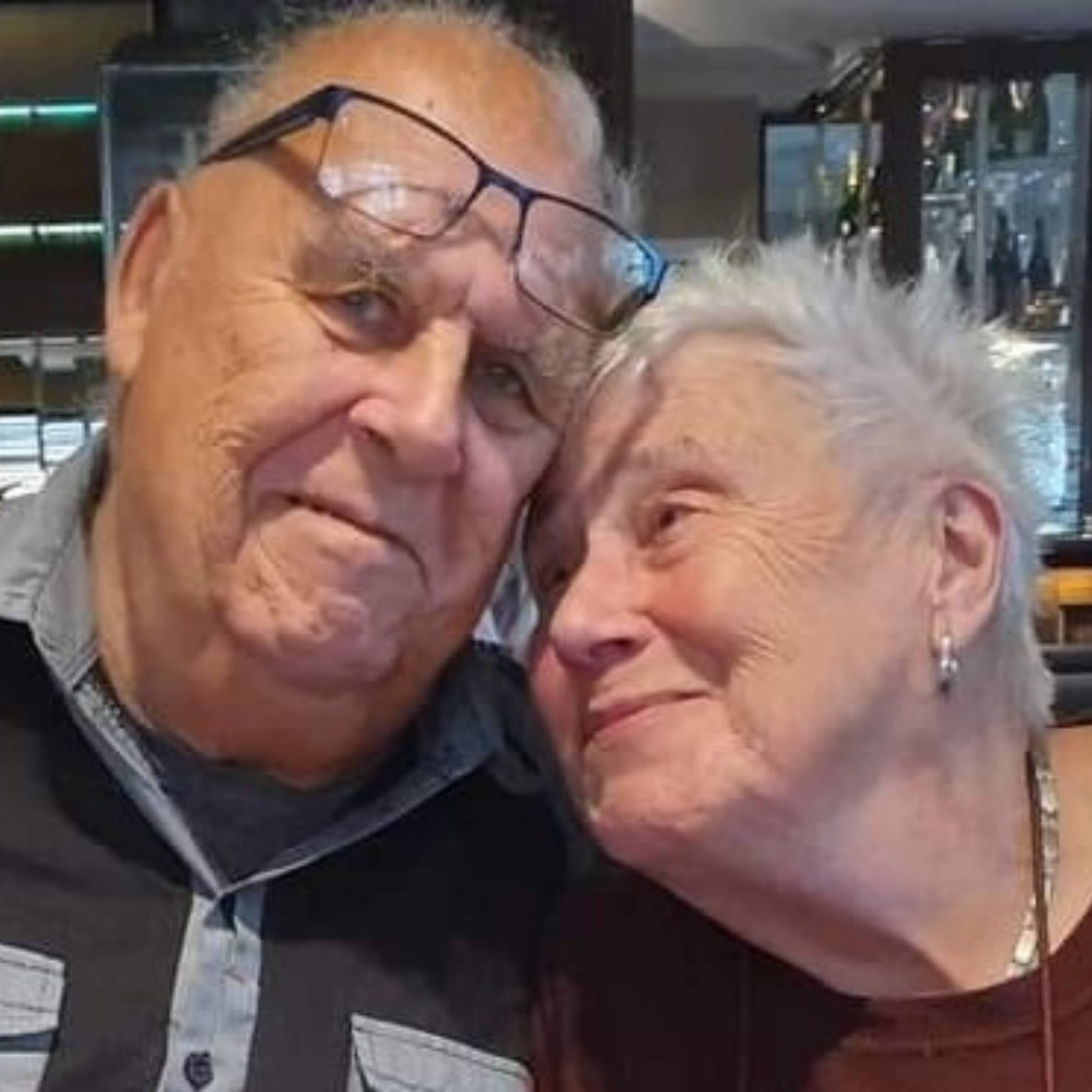 older man wearing glasses and a dark blue shirt next to an older woman with grey hair wearing a red top. There is wine bottles in a bar in the background.