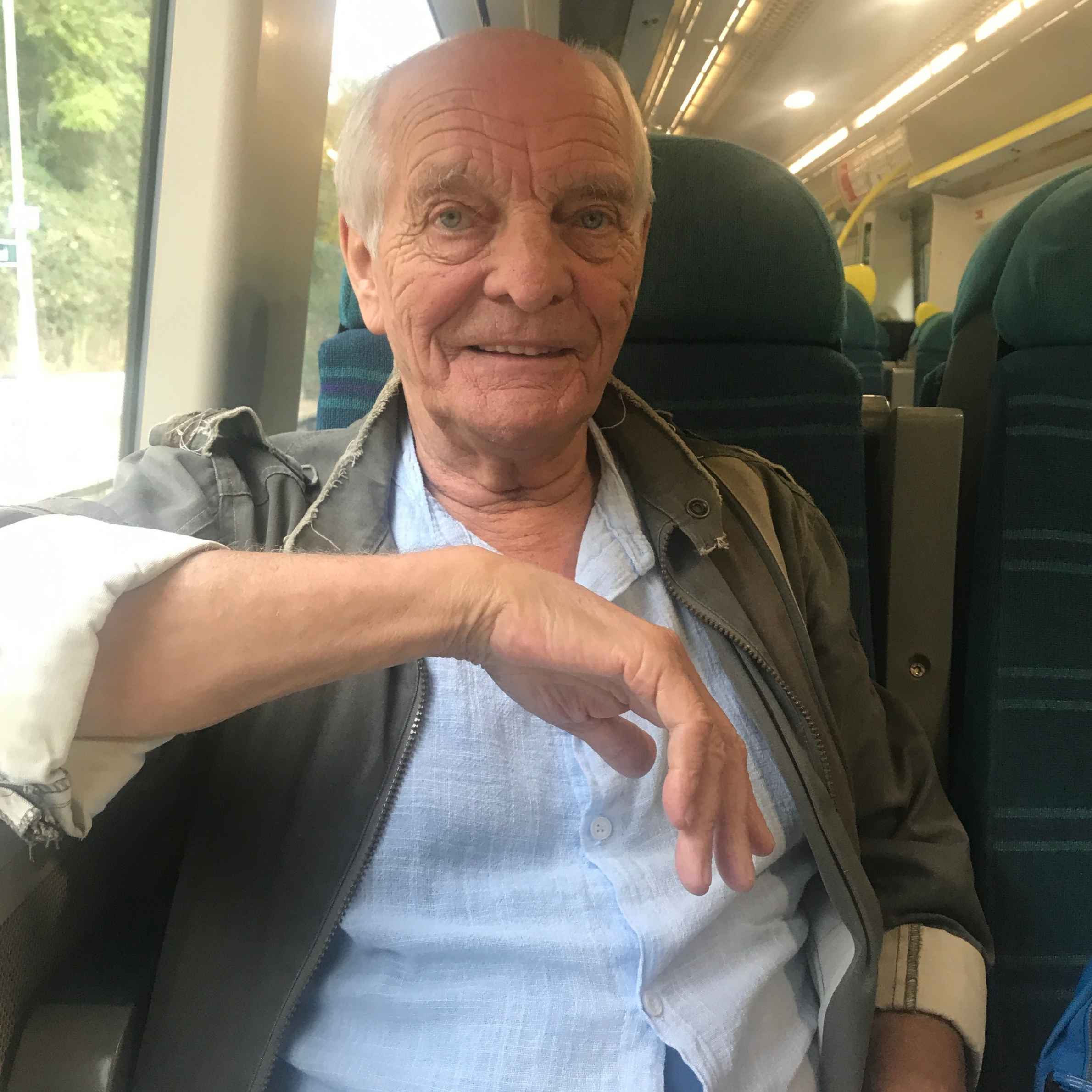 A man sat on the train with blue checked fabric seats. He is wearing a light blue shirt with a jacket over the top. He is smiling, looking face on towards the camera