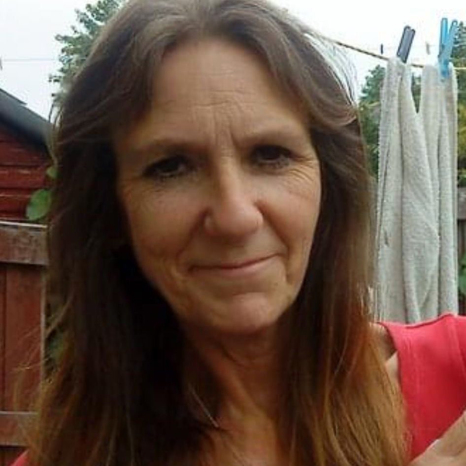 Lady with long brown hair smiling with closed mouth. shes wearing a red top and stood in front of a washing line and brown shed