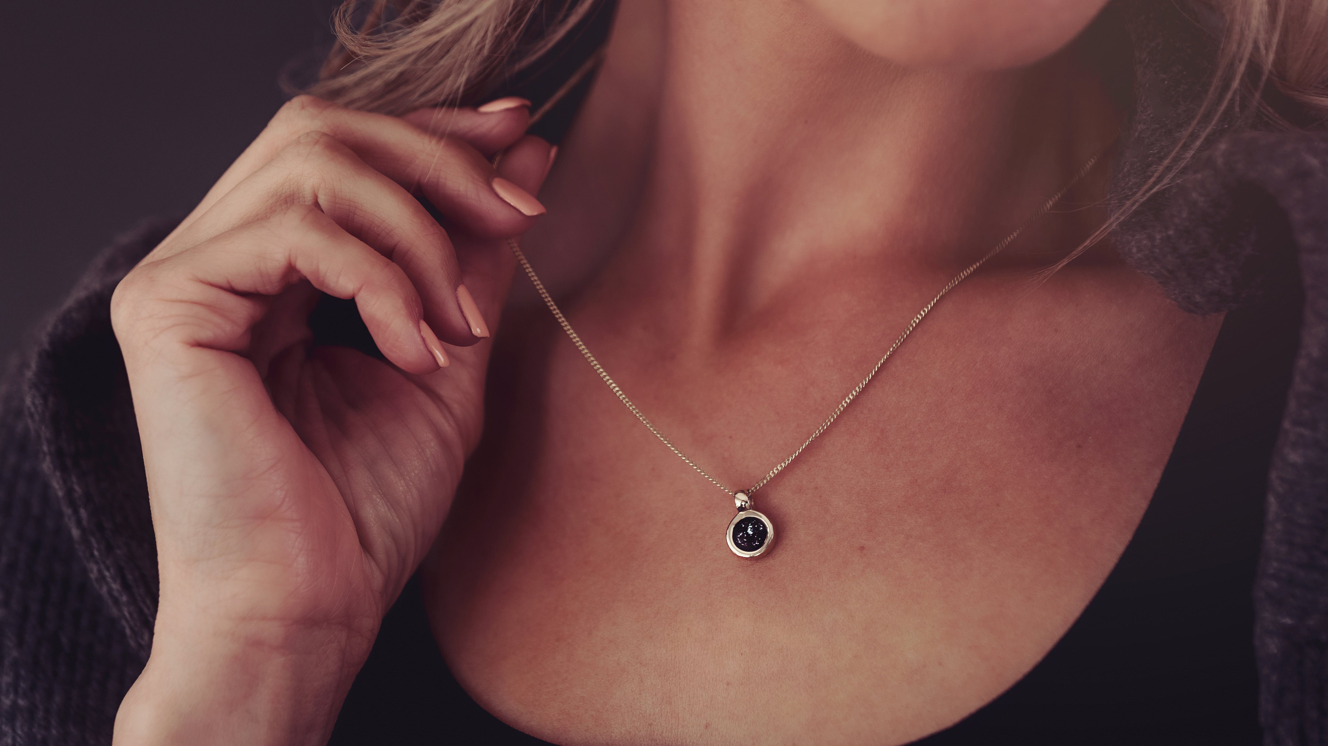 A close-up lifestyle shot of a woman wearing a delicate gold chain necklace with a small round black cremation ashes pendant, set against a dark background.