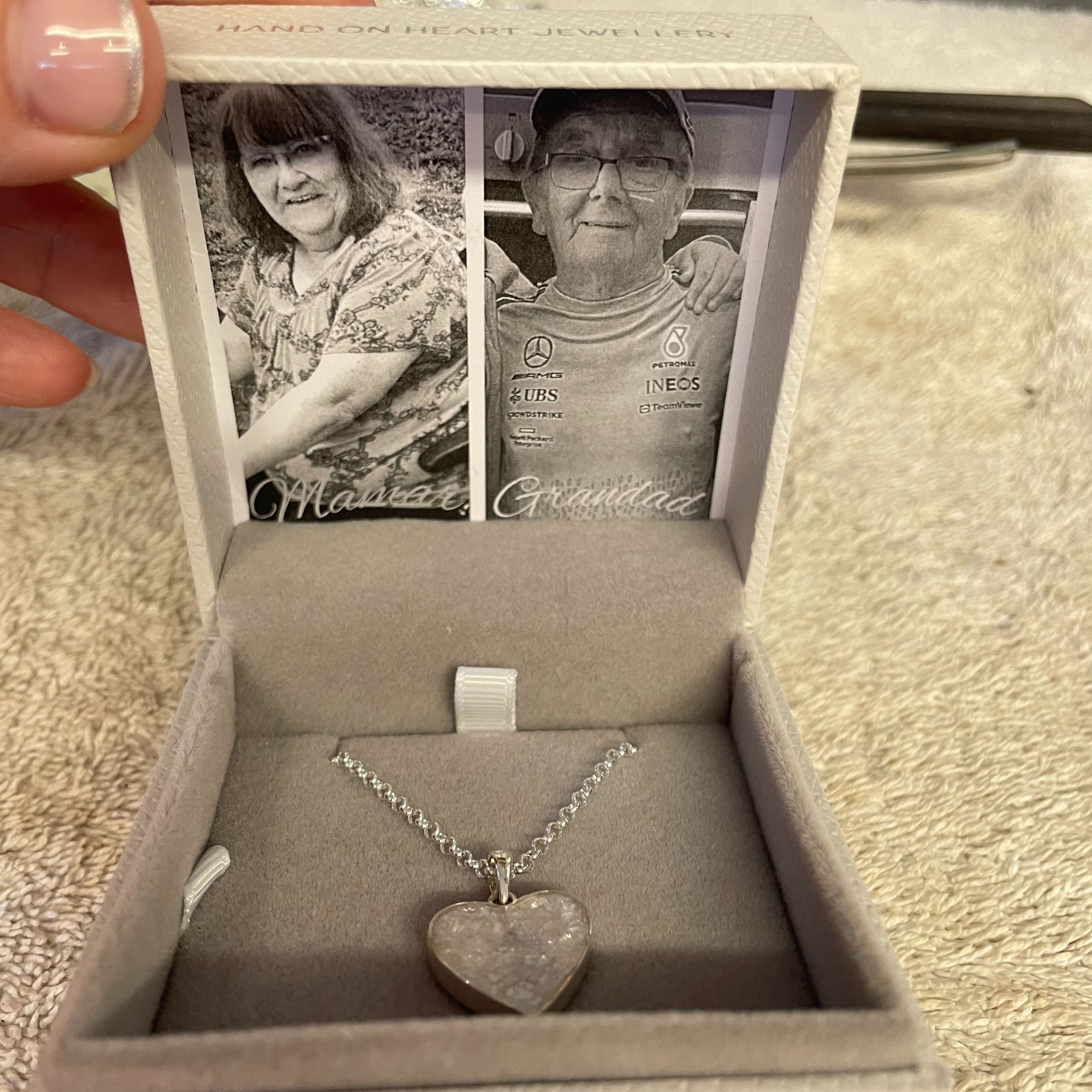 grey jewellery box holding an silver heart necklace with white crystals in it. there is a black and white photo inside the box of a woman and man. the box is on a fluffy grey carpet