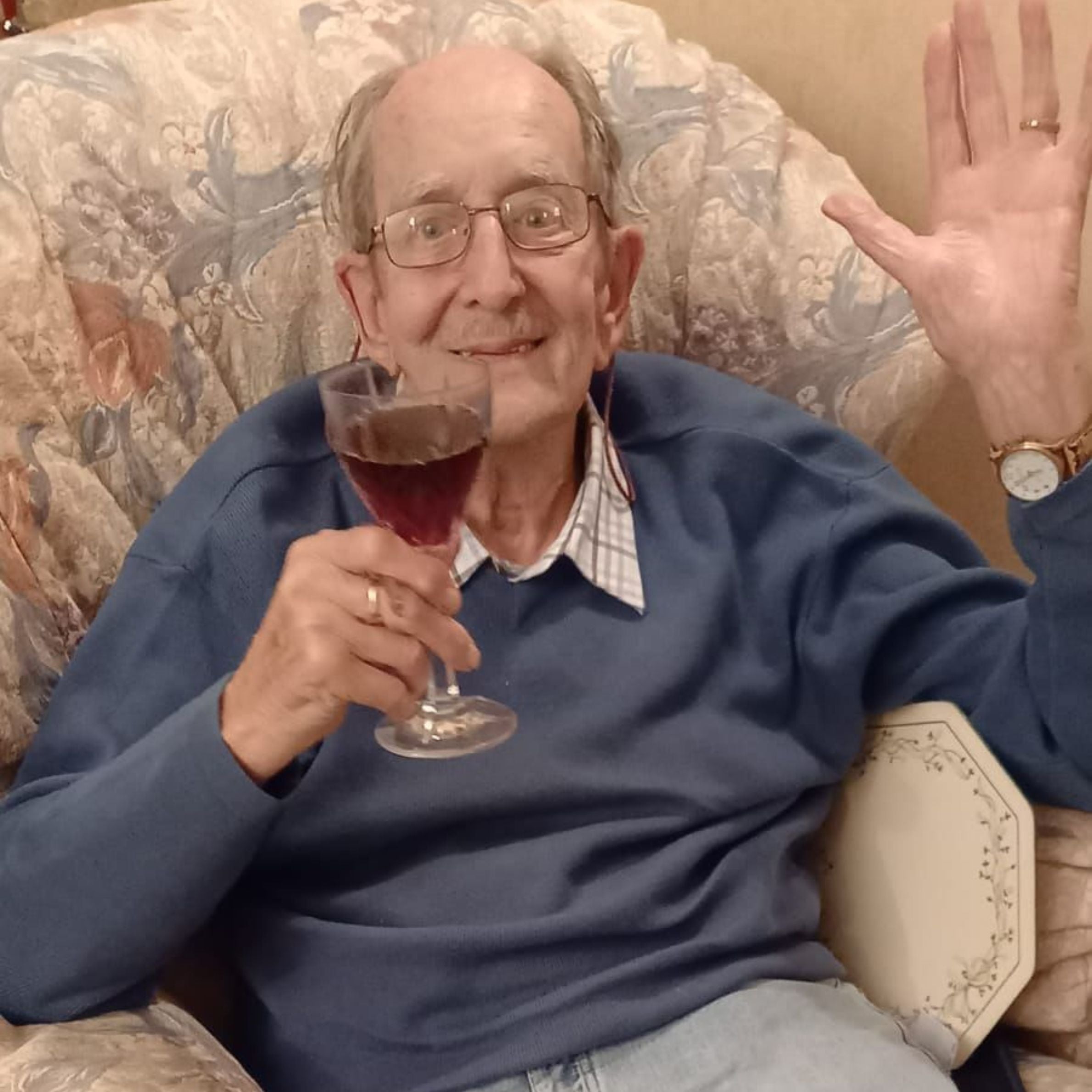 an older man wearing a blue jumper and a pattered collard shirt underneath waving for a photo. he is holding a glass of a red drink and is sat on a beige pattered chair