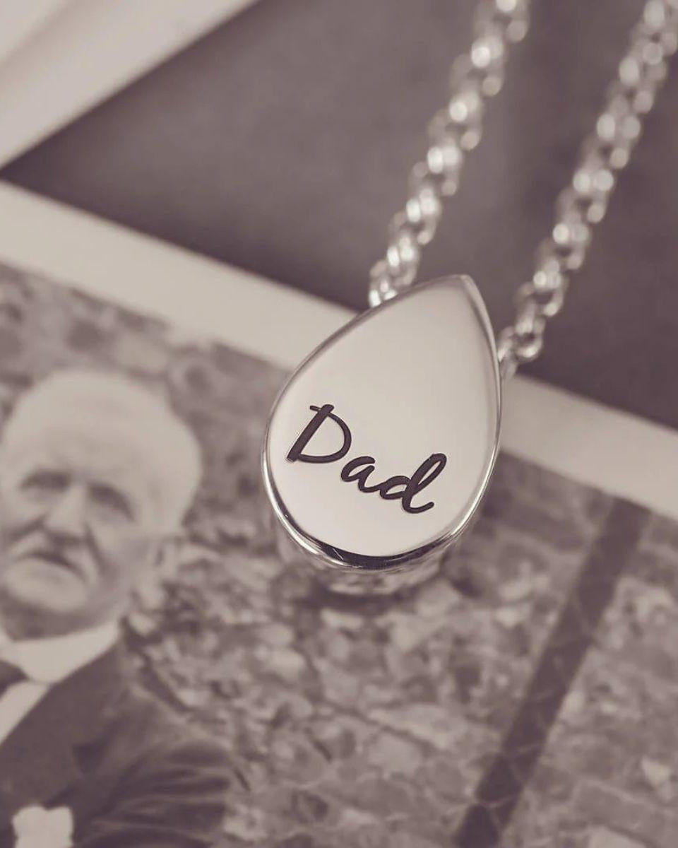 Teardrop-shaped pendant with 'Dad' engraved on it, displayed over a black and white photo of an older man.