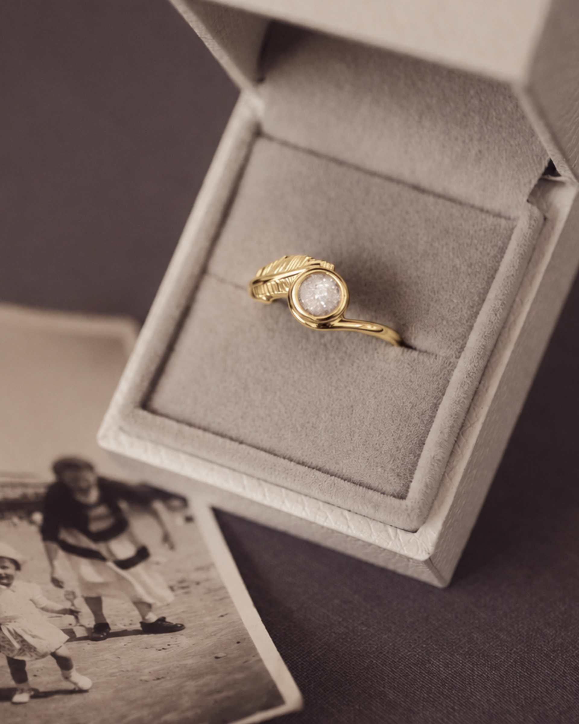 A gold ring with a textured feather detail and a round center stone, displayed inside an open light jewelry box. A vintage-style black-and-white photo lies nearby on a dark surface.