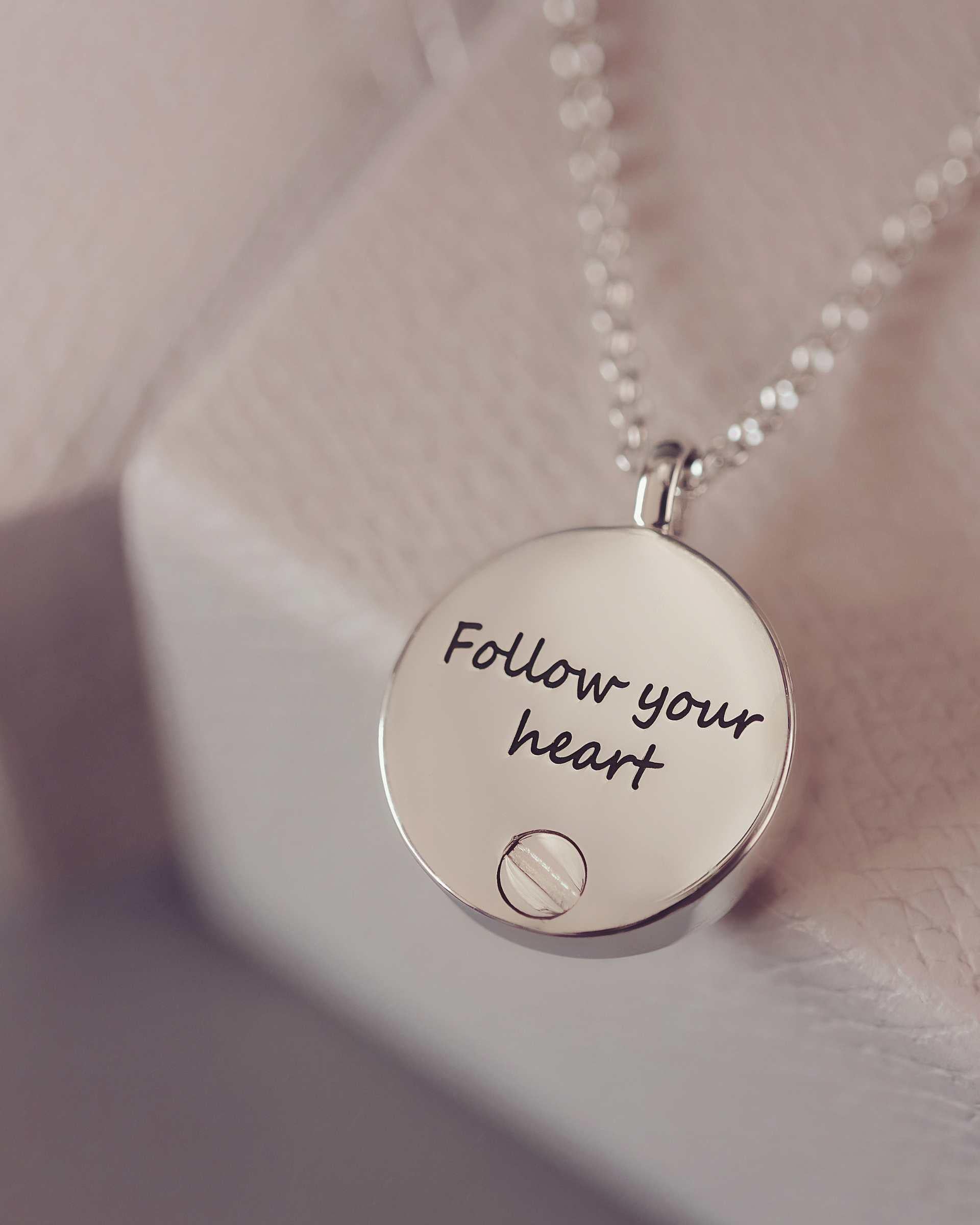 Silver necklace with a round pendant that says 'Follow your heart' on a soft pink background.